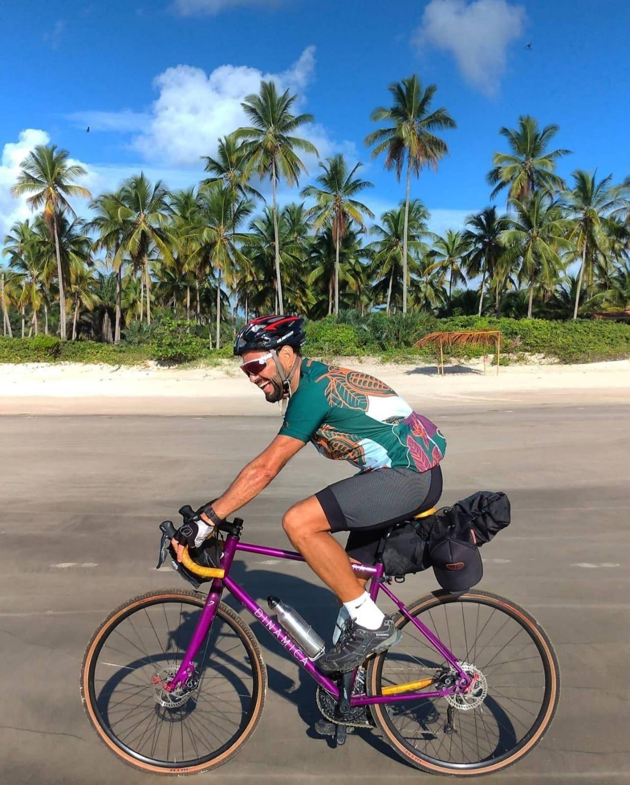 Sun, sand, and open road — the Bahia coastline welcomes every pedal stroke. Smiling cyclist on a loaded purple touring bike riding a coastal road past white sand and tall coconut palms on a cycling Bahia adventure