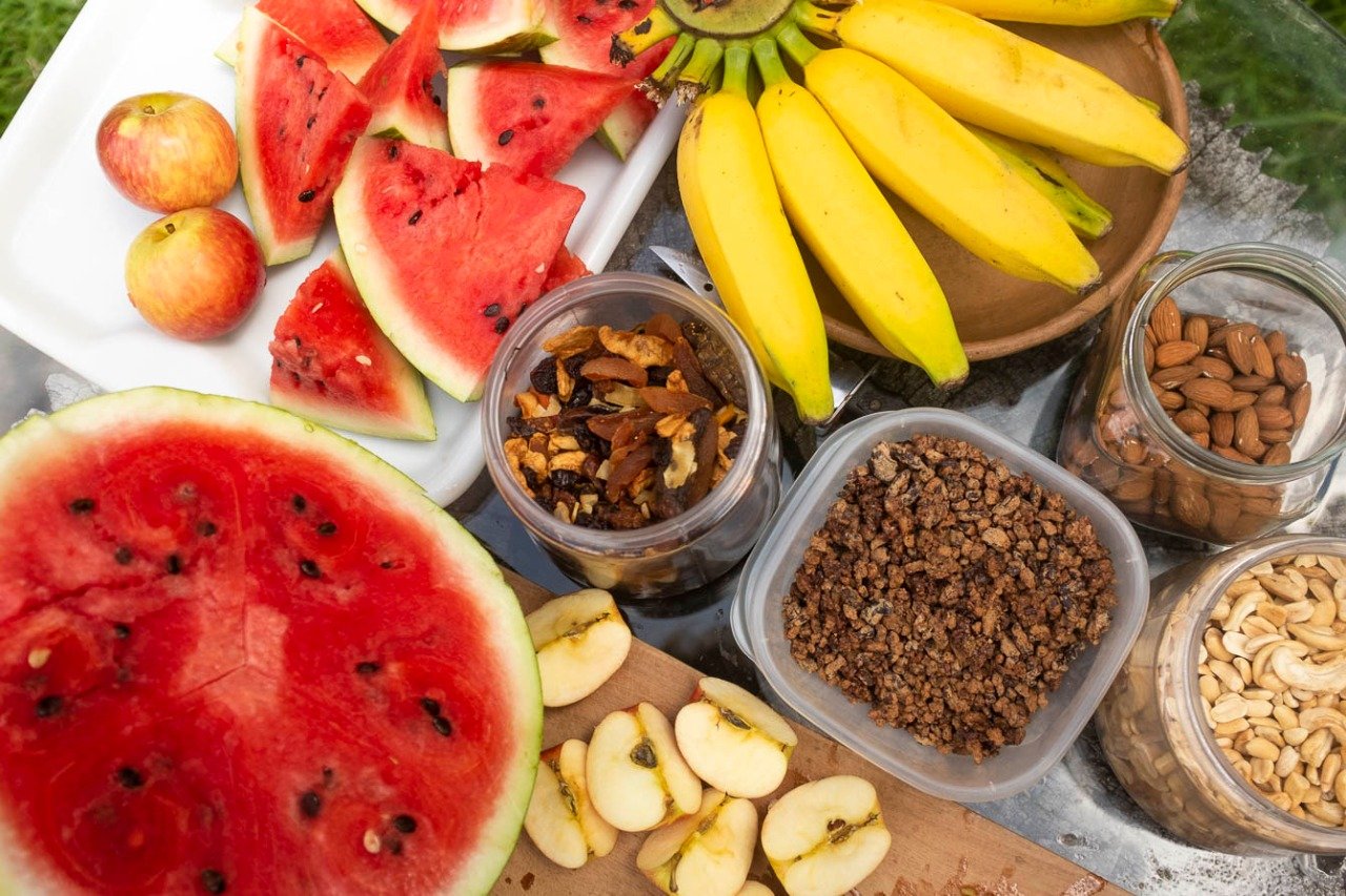 Fuel for the trail — colourful fresh fruit and local nuts ready for the ride Overhead view of a cycling bahia snack spread featuring sliced watermelon, bananas, chopped apples, granola, dried fruit, almonds and cashews arranged on a tray outdoors