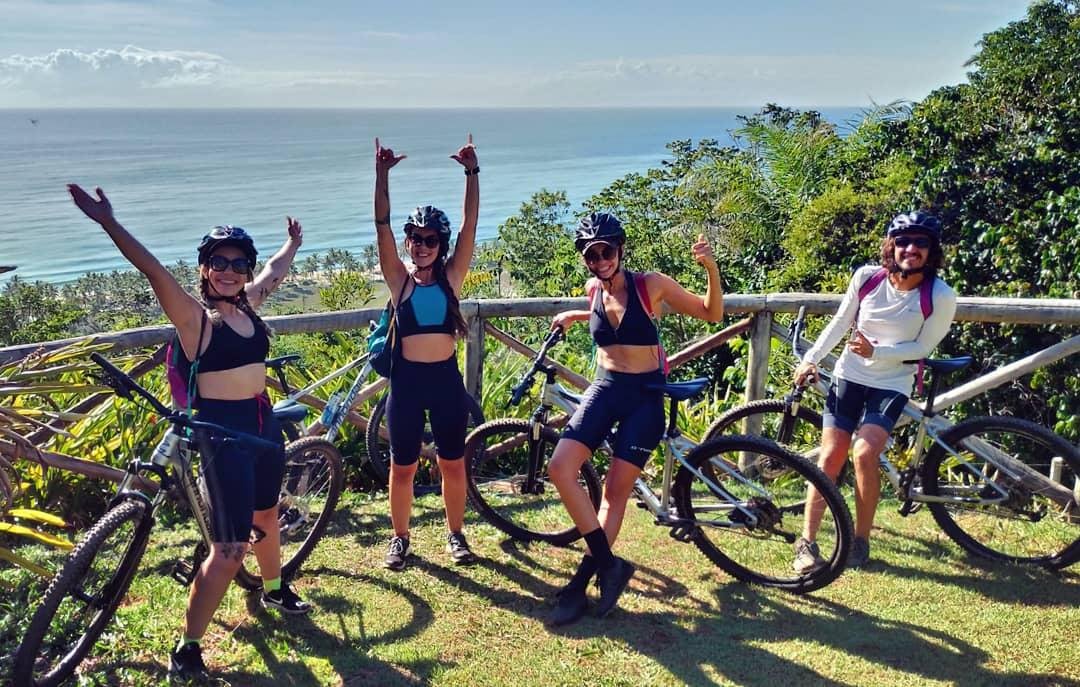 Four cyclists in helmets celebrating at a hilltop viewpoint overlooking Itacaré's turquoise coastline and palm-fringed beach during a cycling Bahia tour