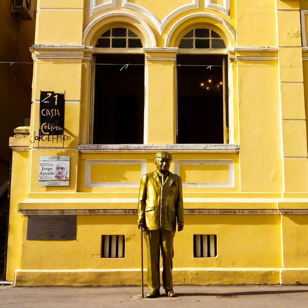 Ilhéus honours its most famous son — the golden Jorge Amado watches over the streets he once wrote about. Golden statue of Jorge Amado standing in front of the bright yellow Casa de Cultura Jorge Amado in Ilhéus, Bahia, a landmark stop on this cycling Bahia experience