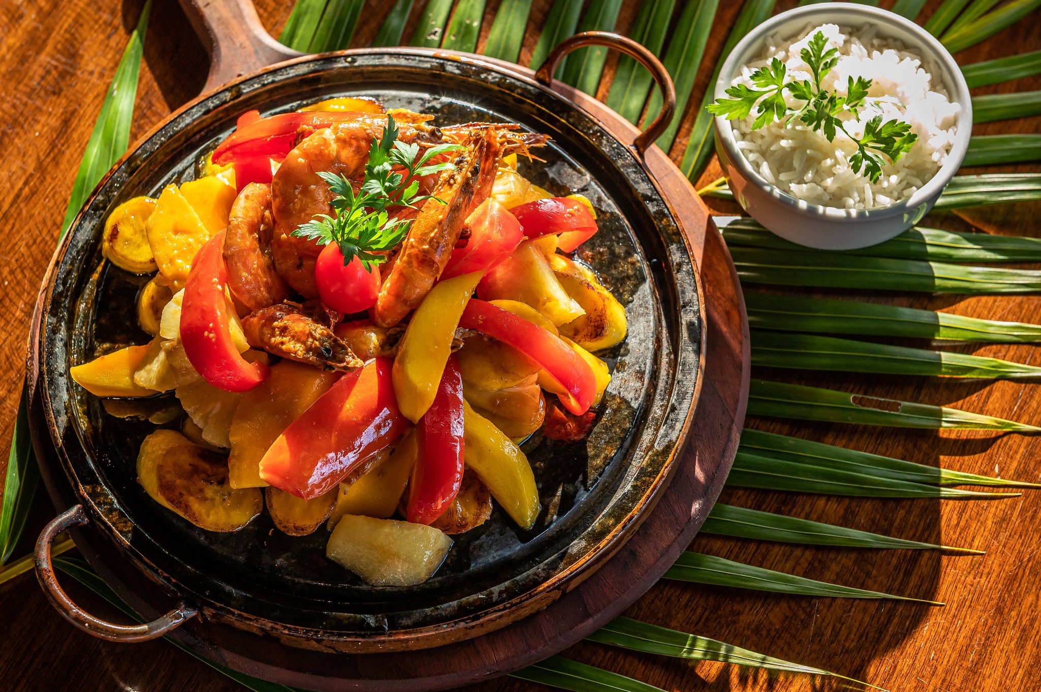 Fresh Atlantic coast flavours — a sizzling prawn skillet waiting at the end of a great ride. Sizzling cast-iron skillet of whole prawns with colourful bell peppers and tropical fruit, served with a bowl of white rice on a palm-leaf-lined wooden board in Itacaré, Bahia