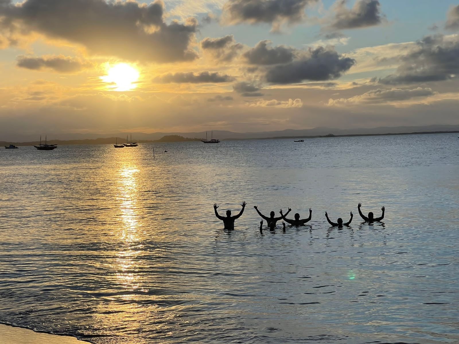 Arms up, sun down — pure joy on the Rio das Contas at golden hour. Five people silhouetted in the calm waters of Rio das Contas at sunset near Itacaré, arms raised in celebration, with sailing boats and distant hills glowing under a golden sky — a highlight of cycling Bahia's Cocoa Route