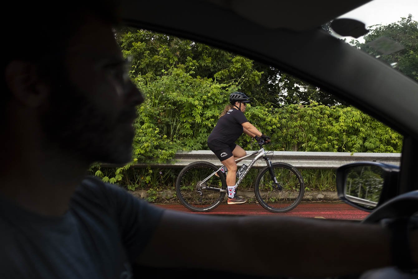 View from inside a support vehicle of a cyclist in black kit and helmet riding a Cannondale mountain bike along a red-surfaced road lined with lush Atlantic Forest near Itacaré, Bahia