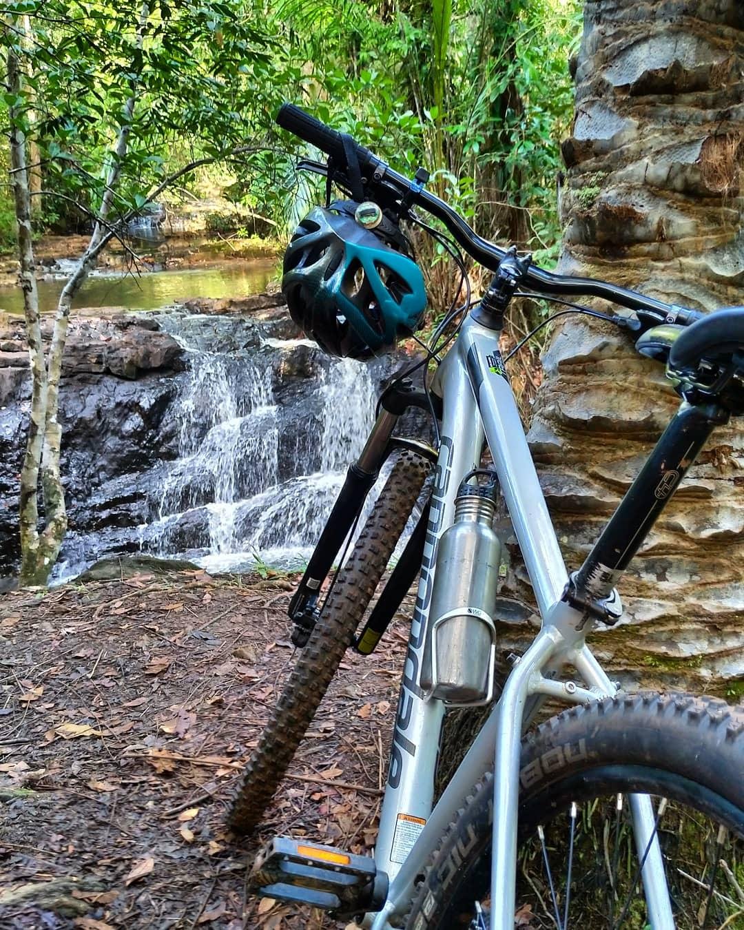 A perfect reason to pause — cool water, ancient rocks, and the trail ahead. Mountain bike with teal helmet resting against a sandstone rock beside a cascading waterfall surrounded by Atlantic Forest on a cycling bahia route near Serra Grande