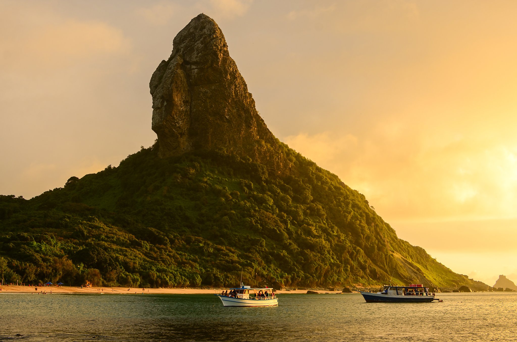 Two boats anchored in calm water at golden sunset in front of the iconic Morro do Pico volcanic peak, Fernando de Noronha, Brazil — a landmark of fernando de noronha diving packages itineraries
