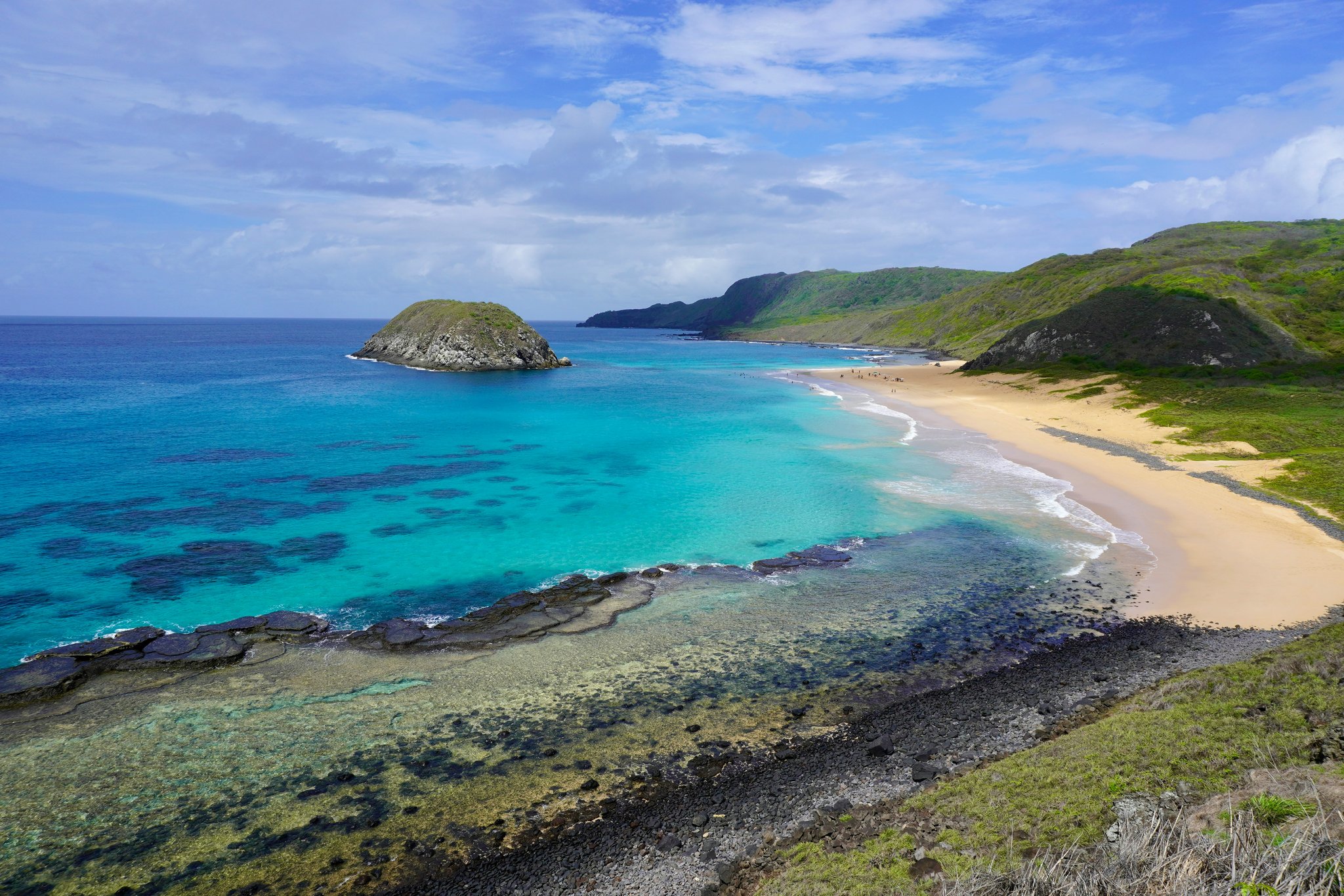 Aerial view of a crescent sandy beach on a Fernando de Noronha diving trip, with turquoise shallow waters revealing coral formations, volcanic rock shelves, a small rocky islet offshore, and green hills framing the bay