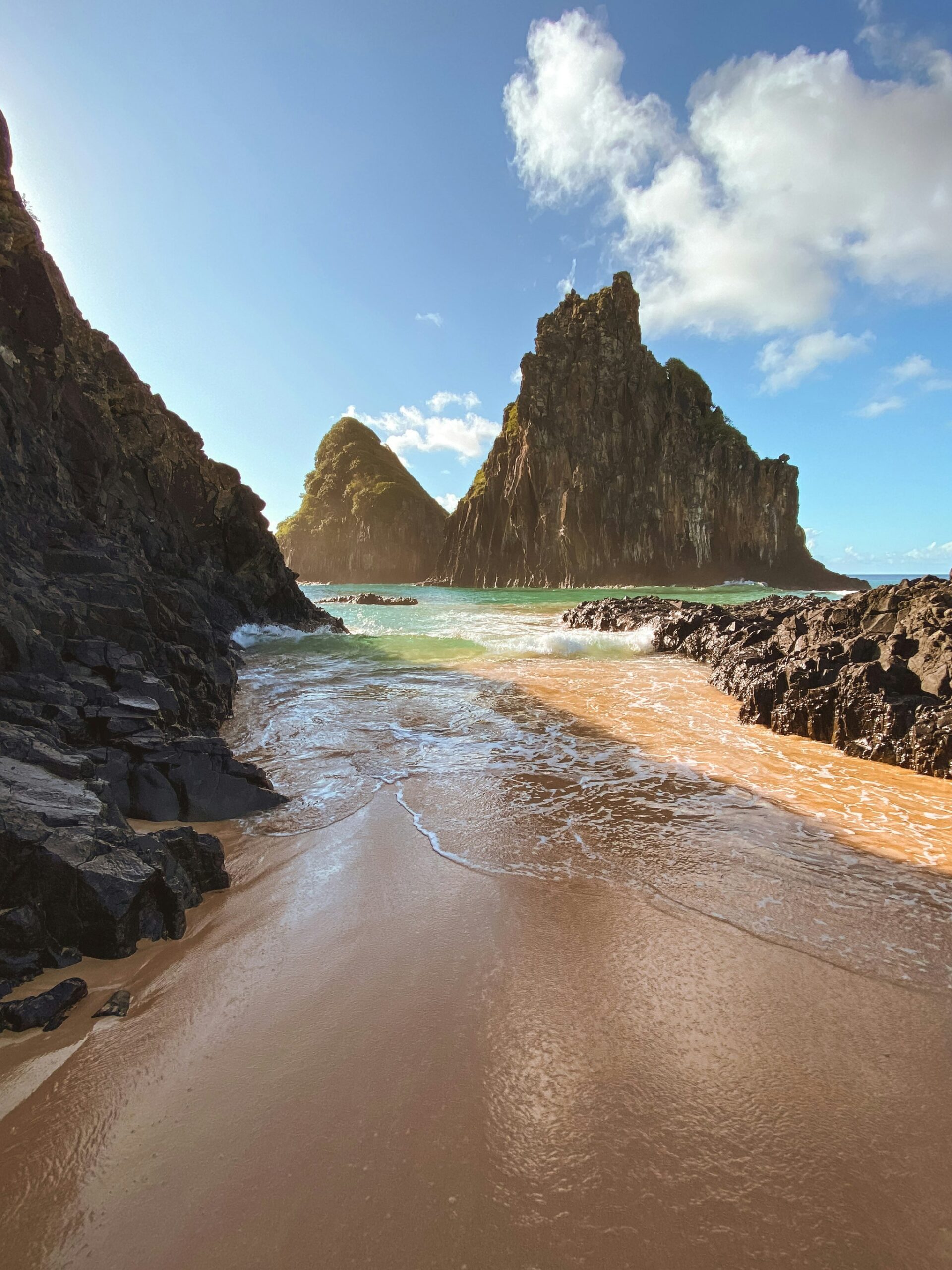 A narrow golden-sand cove flanked by dark volcanic rocks opens onto turquoise Atlantic waters with two towering moss-covered rock spires rising in the background on a Fernando de Noronha diving trip