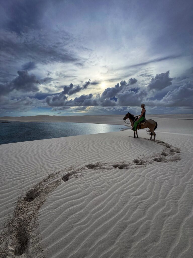 Een soloruiter trekt te paard door de witte zandduinen van Nationaal Park Lençóis Maranhenses, met een regenwaterlagune die zich achter haar uitstrekt.