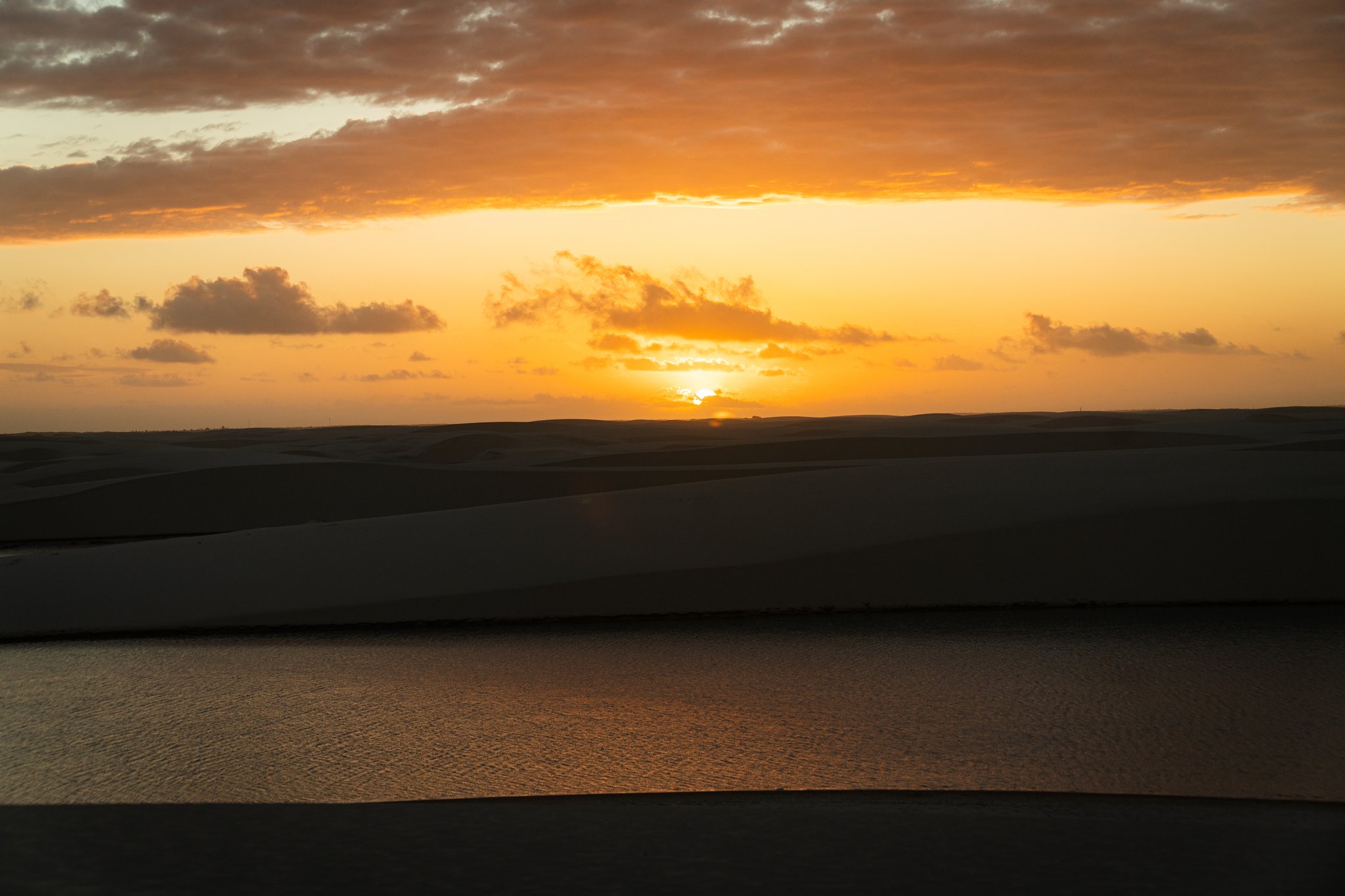 Molten gold poured across the dunes as the sun slips below the horizon. Golden sunset over silhouetted sand dunes and a still lagoon reflecting amber light on the horseback riding Lençóis Maranhenses tour
