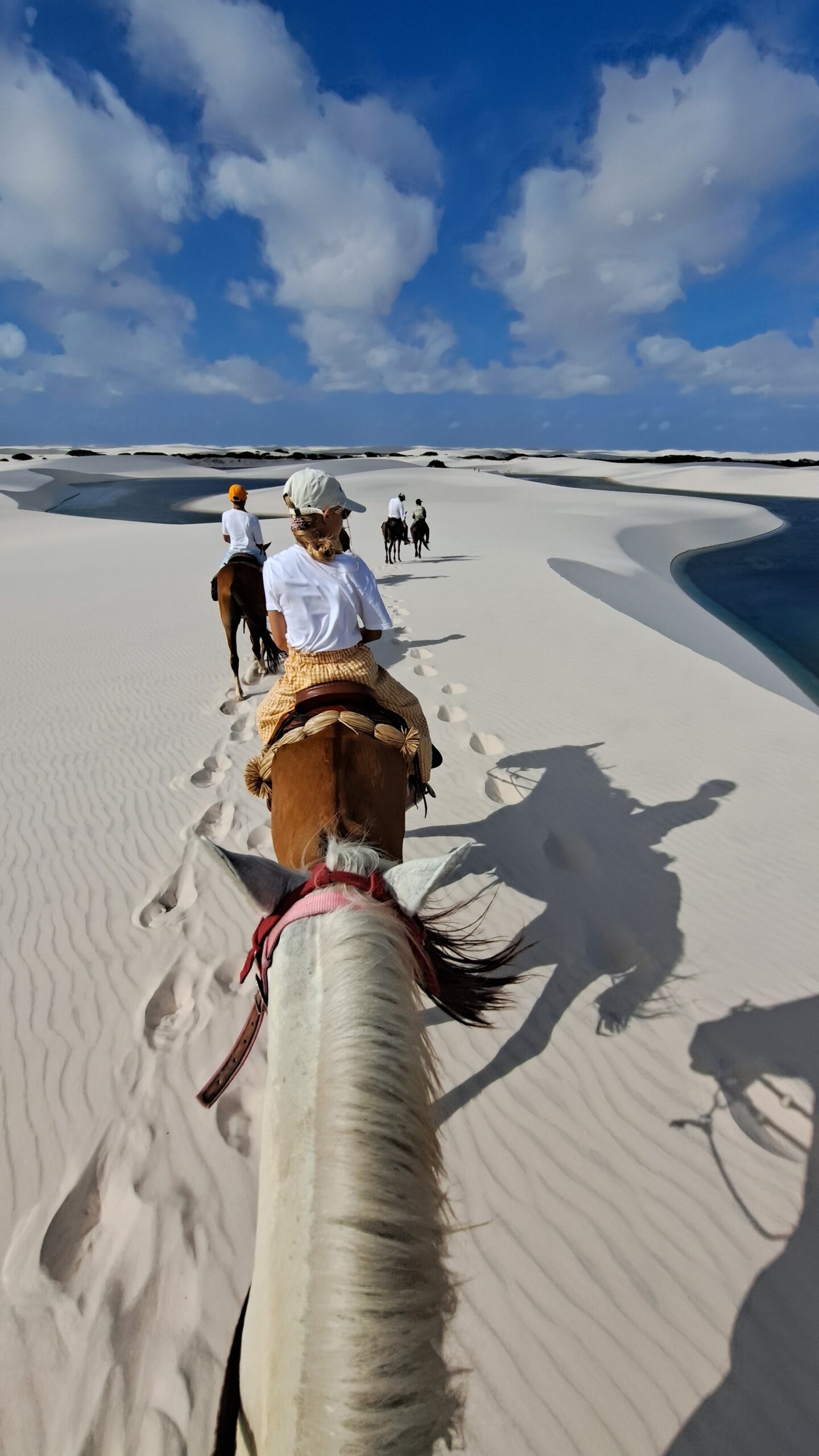 Hoofprints in the sand, lagoons shimmering ahead — this is how you cross Lençóis Maranhenses. Point-of-view shot from horseback on a horseback riding Lençóis Maranhenses tour, with riders and hoofprints tracing a line across rippled white dunes beside a turquoise lagoon under a bright blue sky