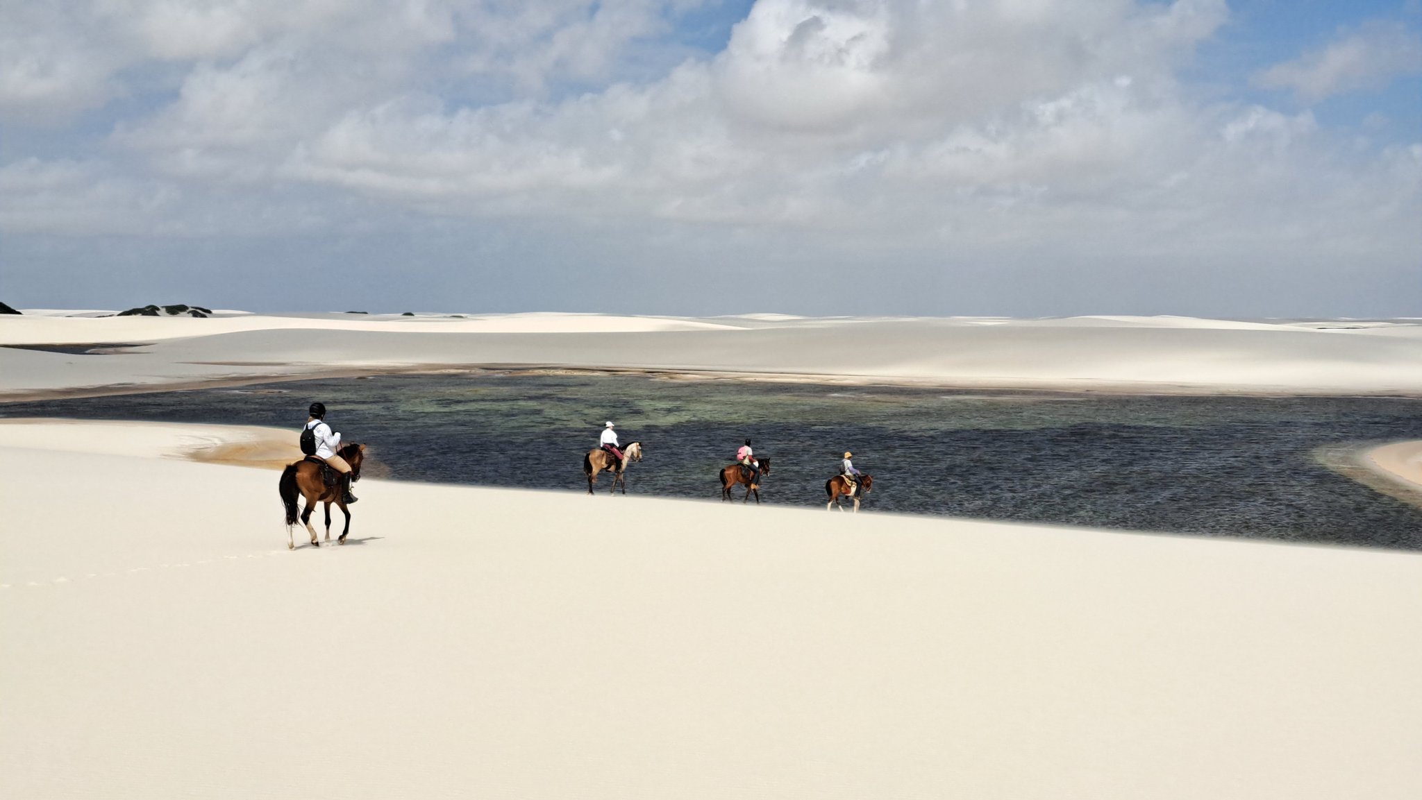 Four horses, one sweeping lagoon — the dunes stretch on forever. Four riders on horseback cross white sand dunes beside a dark freshwater lagoon on a horseback riding Lençóis Maranhenses tour, under a partly cloudy sky