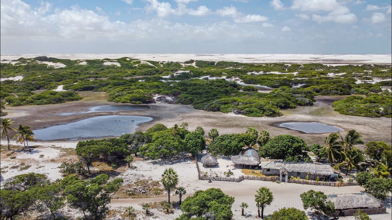 Tiny thatched homes tucked between dunes and lagoons — life at the heart of the park. Aerial view of a small thatched-roof village nestled among white sand dunes, scrub vegetation and shallow lagoons on the horseback riding Lençóis Maranhenses tour route