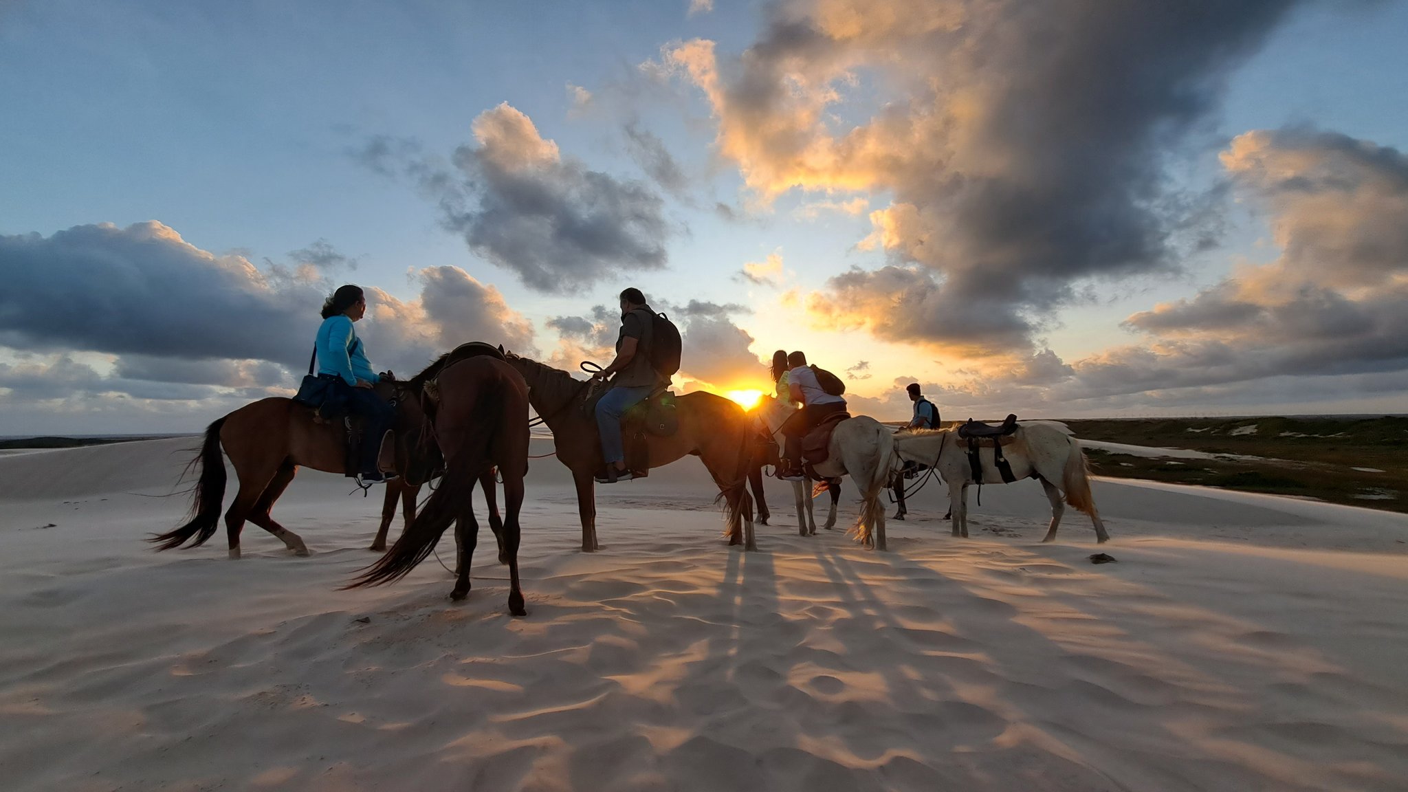 Riders paused at golden hour, shadows stretching long across the dunes. A group of riders on horseback silhouetted against a blazing sunset over the white sand dunes of Lençóis Maranhenses, with dramatic clouds spreading across the sky during a dune crossing on horseback
