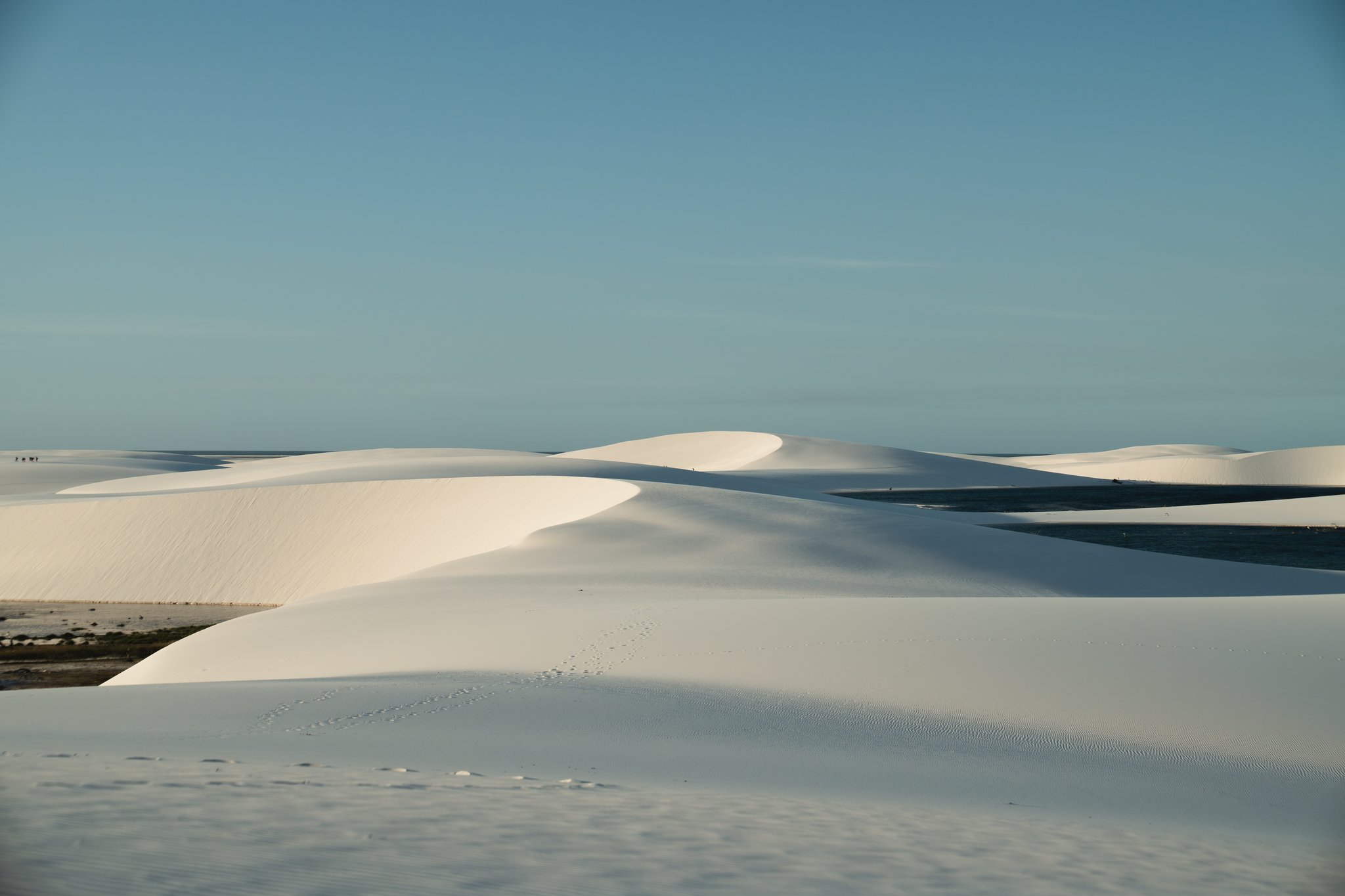 Rippled white silk — dune after dune, as far as the eye can reach. Sweeping white sand dunes of Lençóis Maranhenses stretch to the horizon beneath a clear blue sky, with shallow lagoons visible between the sculpted ridges on the horseback riding Lençóis Maranhenses tour route