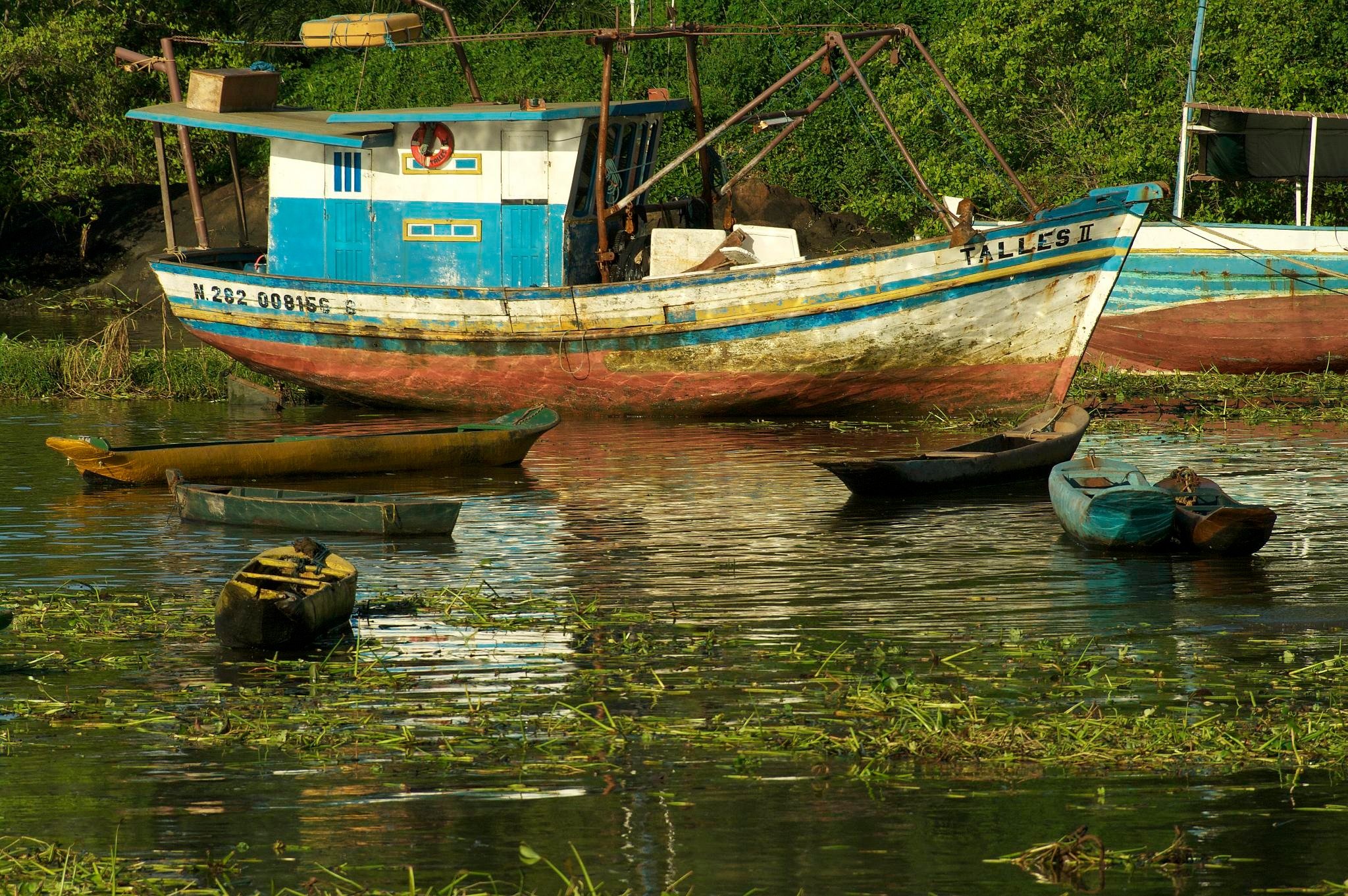 Weathered blue-and-white fishing boat named Talles II moored alongside small wooden canoes on a vegetation-lined river in Itacaré, Bahia