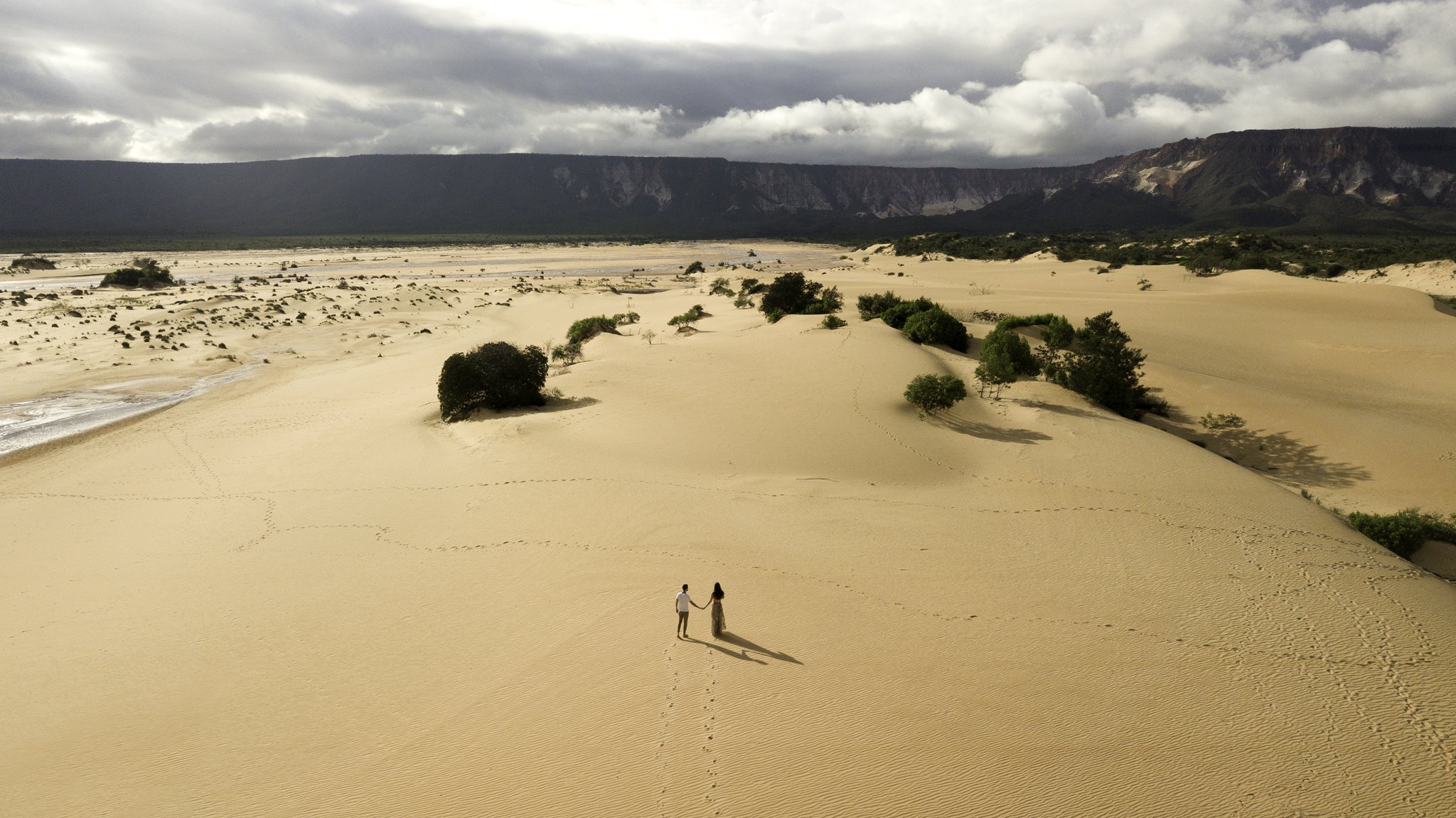 Tiny figures, endless dunes — footprints the only mark on the sand. Photo: FG Trade Aerial view of a couple holding hands on the golden Jalapão sand dunes, with scattered green shrubs, a river edge and dramatic mesa cliffs under a stormy sky during a Jalapão adventure