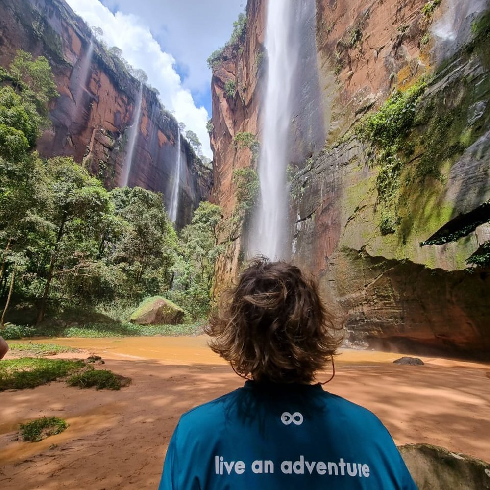 Waterfalls pouring off ancient sandstone, all at once. A traveler in a teal 'live an adventure' shirt gazes up at multiple waterfalls cascading down towering red sandstone cliffs into an ochre-tinted pool — a defining moment on a Jalapão adventure