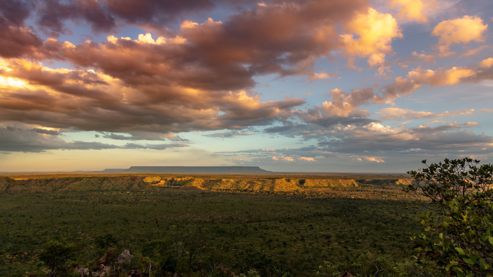 A vast Cerrado landscape at sunset during a Jalapão adventure, with a flat-topped mesa silhouetted against a dramatic sky of golden and rose-tinted clouds