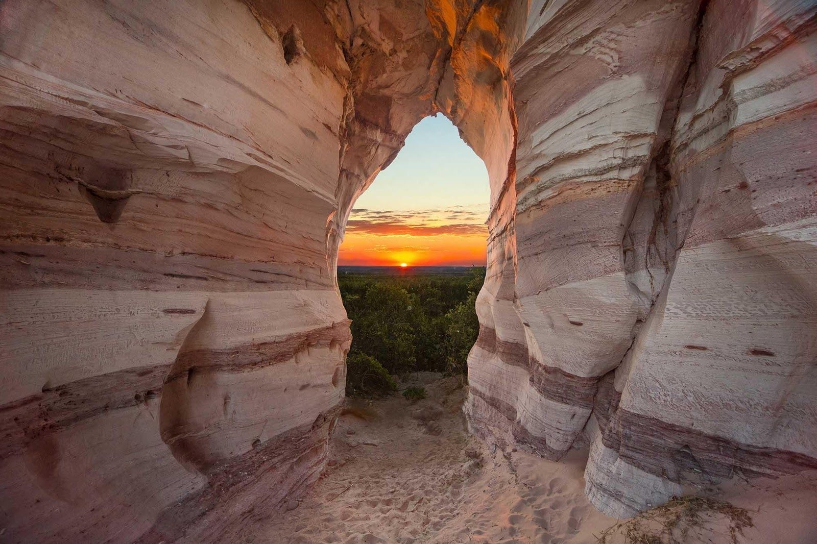 Sunset framed perfectly — the ancient stone arch of Pedra Furada holds a burning horizon. Warm light glows through a natural arch in layered sandstone rock during a jalapão adventure, framing a fiery sunset over a vast green canopy below