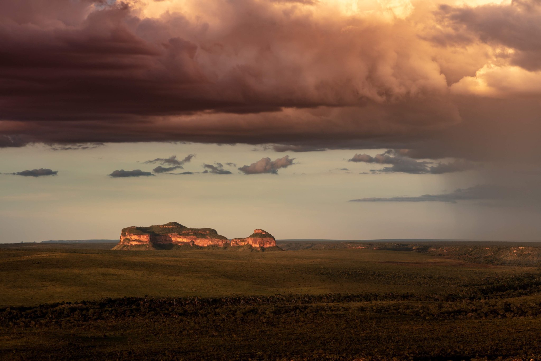 Two flat-topped sandstone mesas glow amber in last light beneath a dramatic storm front rolling across an open Cerrado plain on a Jalapão adventure