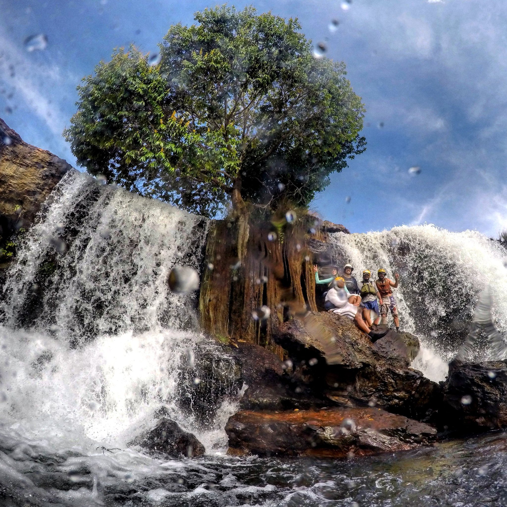 Helmets on, spray everywhere — wedged between twin falls and loving every second. A group of travellers in safety helmets and life jackets perched on rocky outcrops between two powerful cascading waterfalls during a Jalapão adventure, with mist and spray filling the air and a lone tree rising above the falls against a blue sky