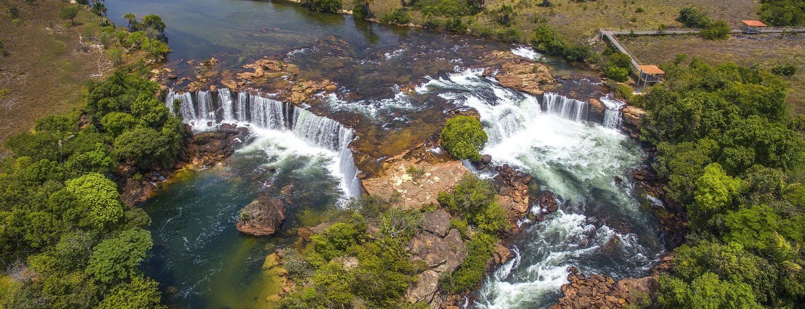 Aerial view of a wide multi-tiered waterfall cascading over flat sandstone rocks into green pools, surrounded by Cerrado vegetation in Jalapão, Brazil