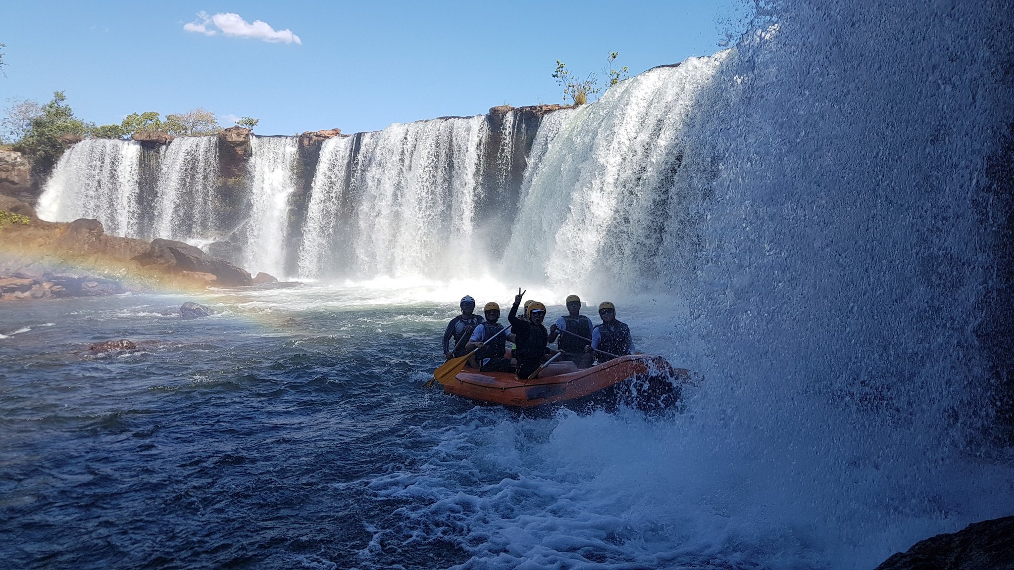 A group of helmeted rafters in an orange inflatable raft paddling through the powerful spray at the base of a wide Jalapão waterfall, with a rainbow visible in the mist