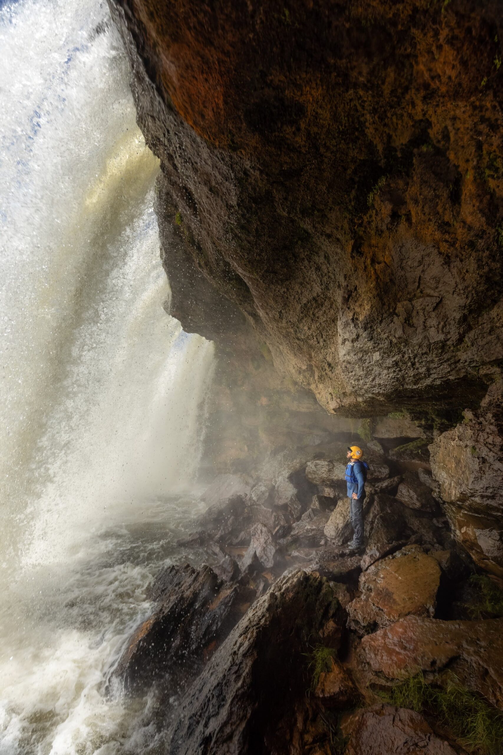 A hiker in a blue jacket and orange helmet stands on a rocky ledge beneath a massive thundering waterfall during a Jalapão trekking expedition, dwarfed by the cascading white water and overhanging sandstone cliff
