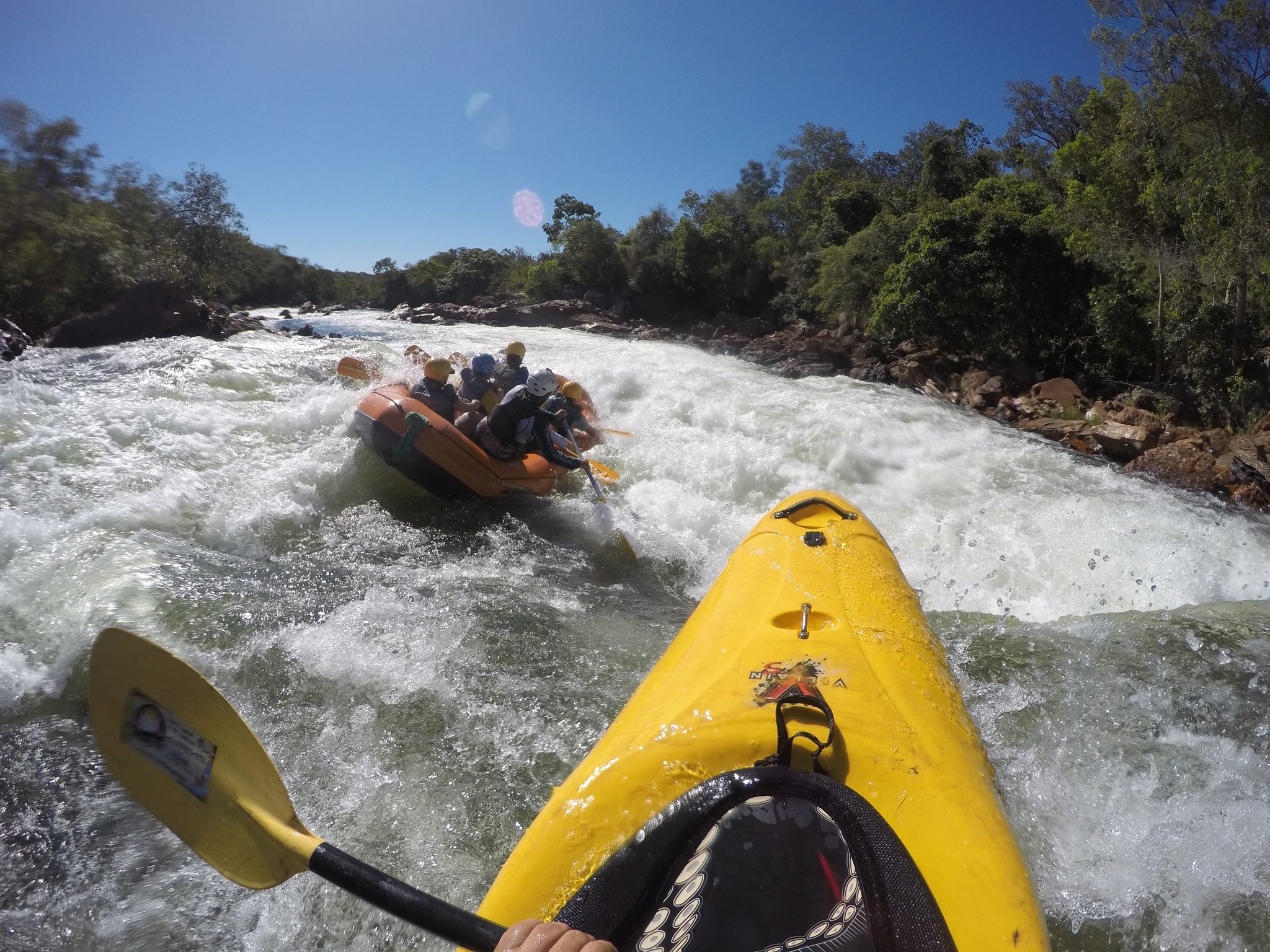 POV from a yellow kayak on churning white-water rapids as a crew paddles an orange raft through a rocky gorge lined with green trees during Jalapão trekking