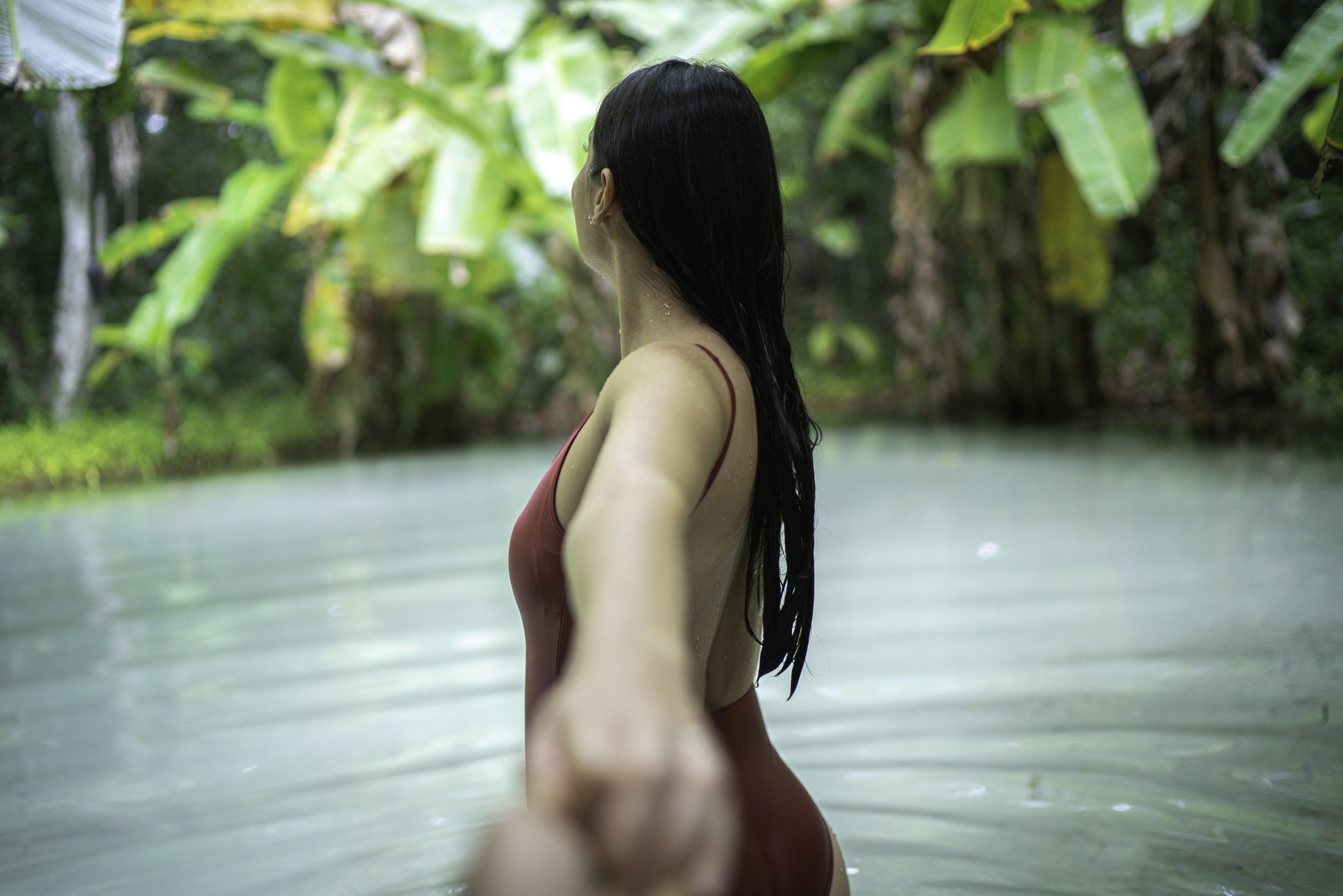 A woman in a red swimsuit wades through a pale turquoise natural spring pool surrounded by lush tropical vegetation, reaching back toward the camera during a Jalapão trekking experience