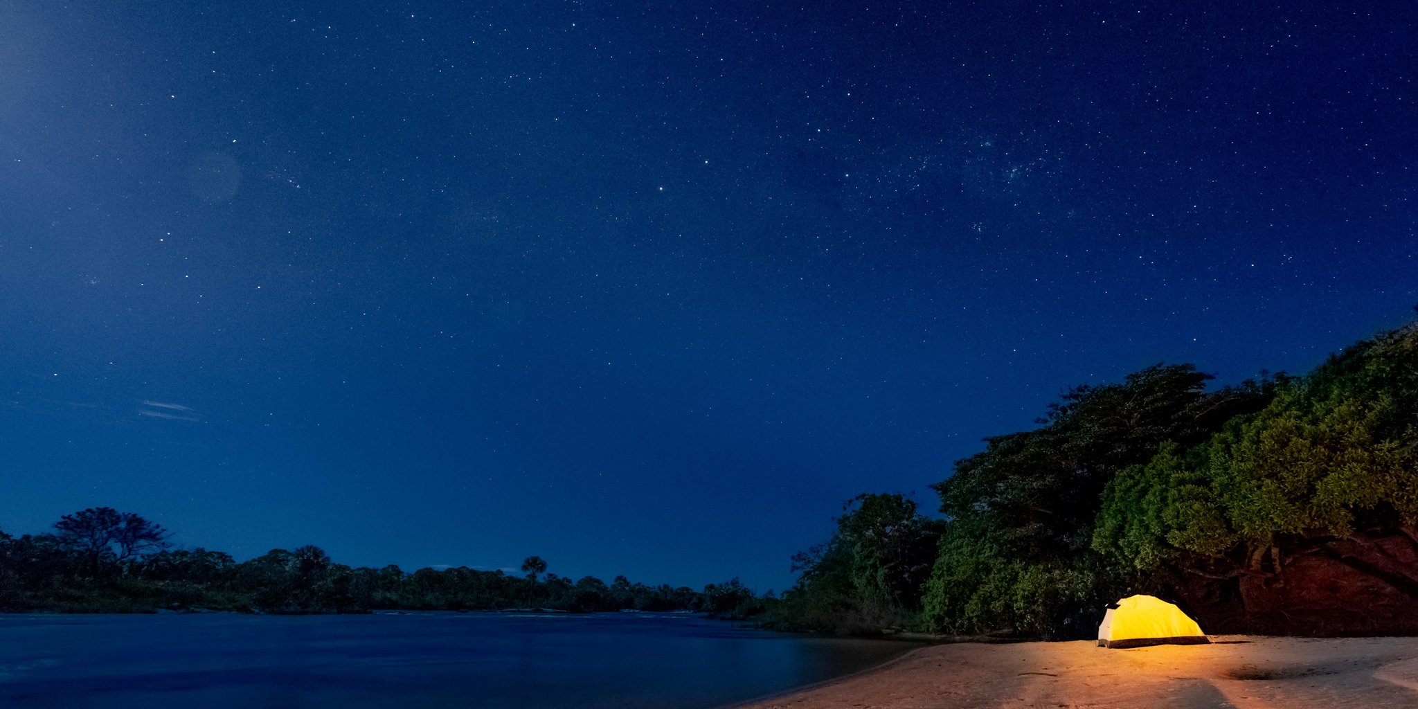 A single illuminated yellow tent pitched on a sandy river beach during a Jalapão trekking expedition, surrounded by dense tropical vegetation under a vast star-filled night sky