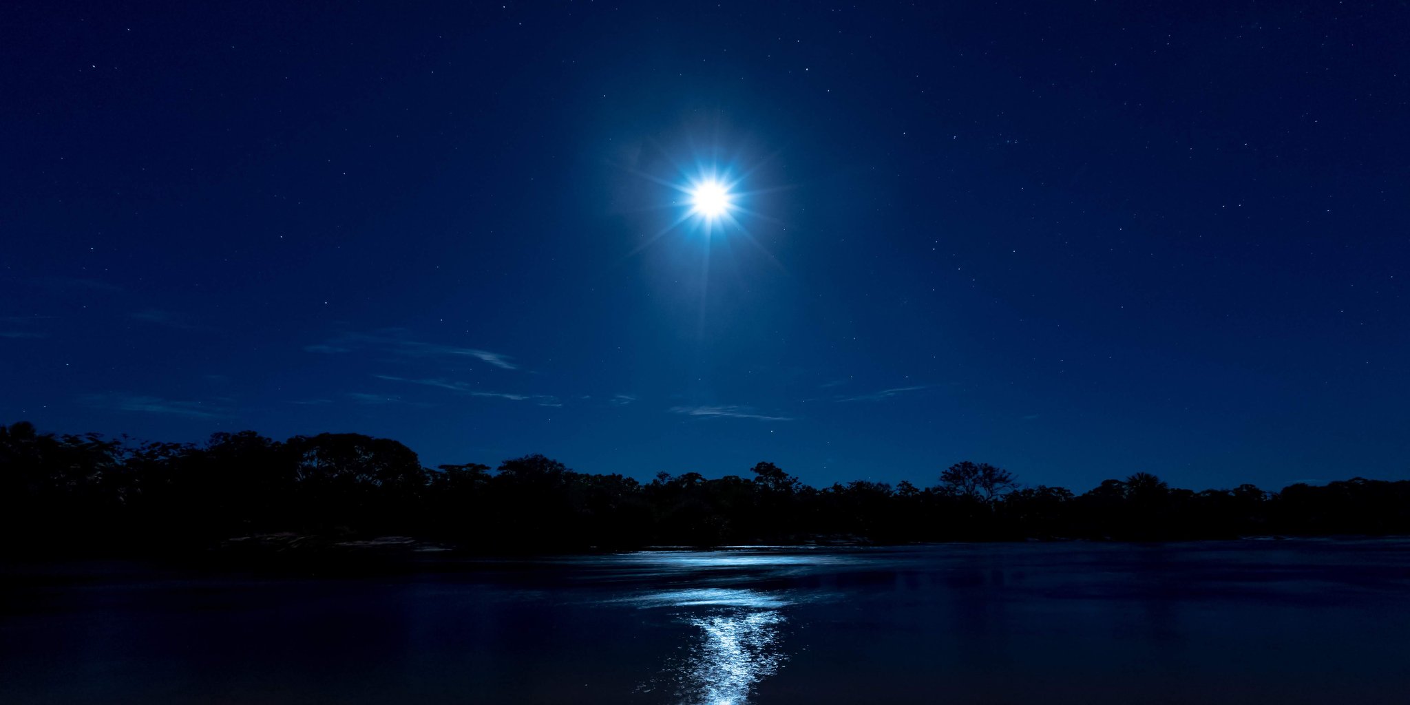 A brilliant full moon rises over a calm river on a Jalapão trekking expedition, its reflection shimmering across the dark water beneath a star-filled deep-blue sky and silhouetted tree line