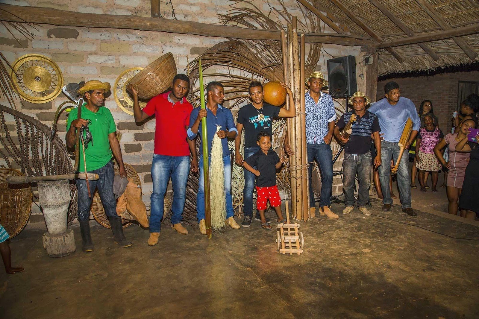 Local men and a child inside a thatched community hall display traditional handcrafted objects — woven baskets, a gourd, carved wood tools, and golden-grass items — during a Jalapão trekking cultural visit to a quilombola community