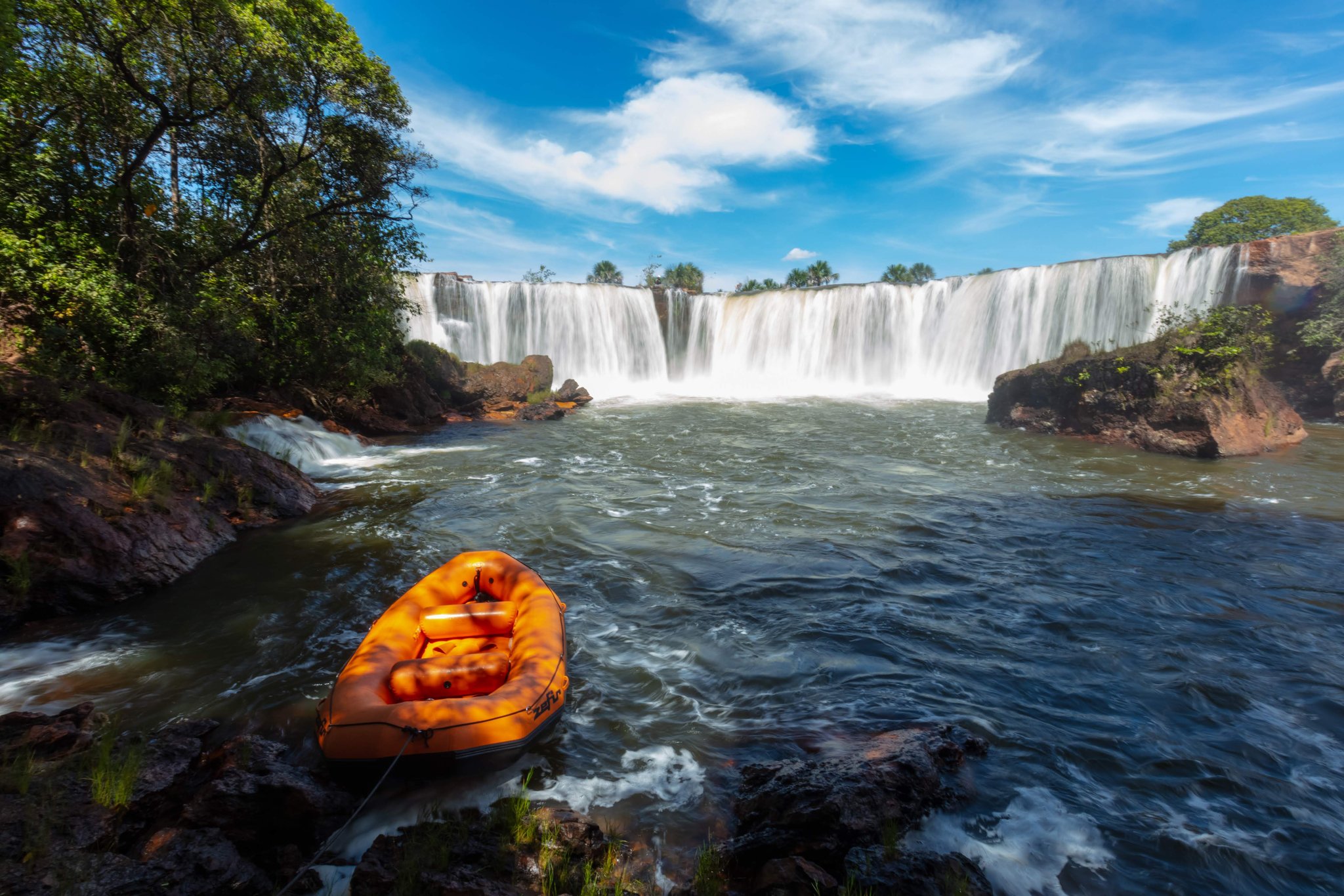 An orange inflatable raft moored on rocky shoreline in front of a wide horseshoe-shaped waterfall cascading into a churning pool on a Jalapão trekking expedition