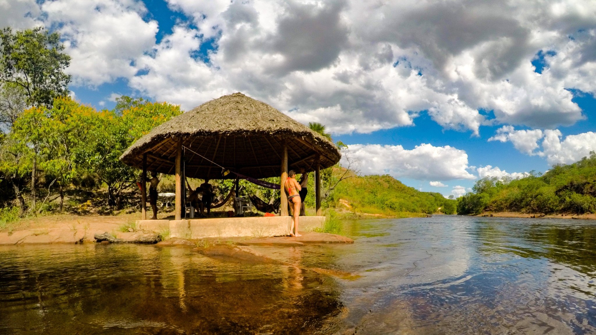 Travelers relaxing under a round thatched-roof shelter on the banks of a clear river in Jalapão trekking country, with hammocks strung inside and green cerrado hills rising in the background