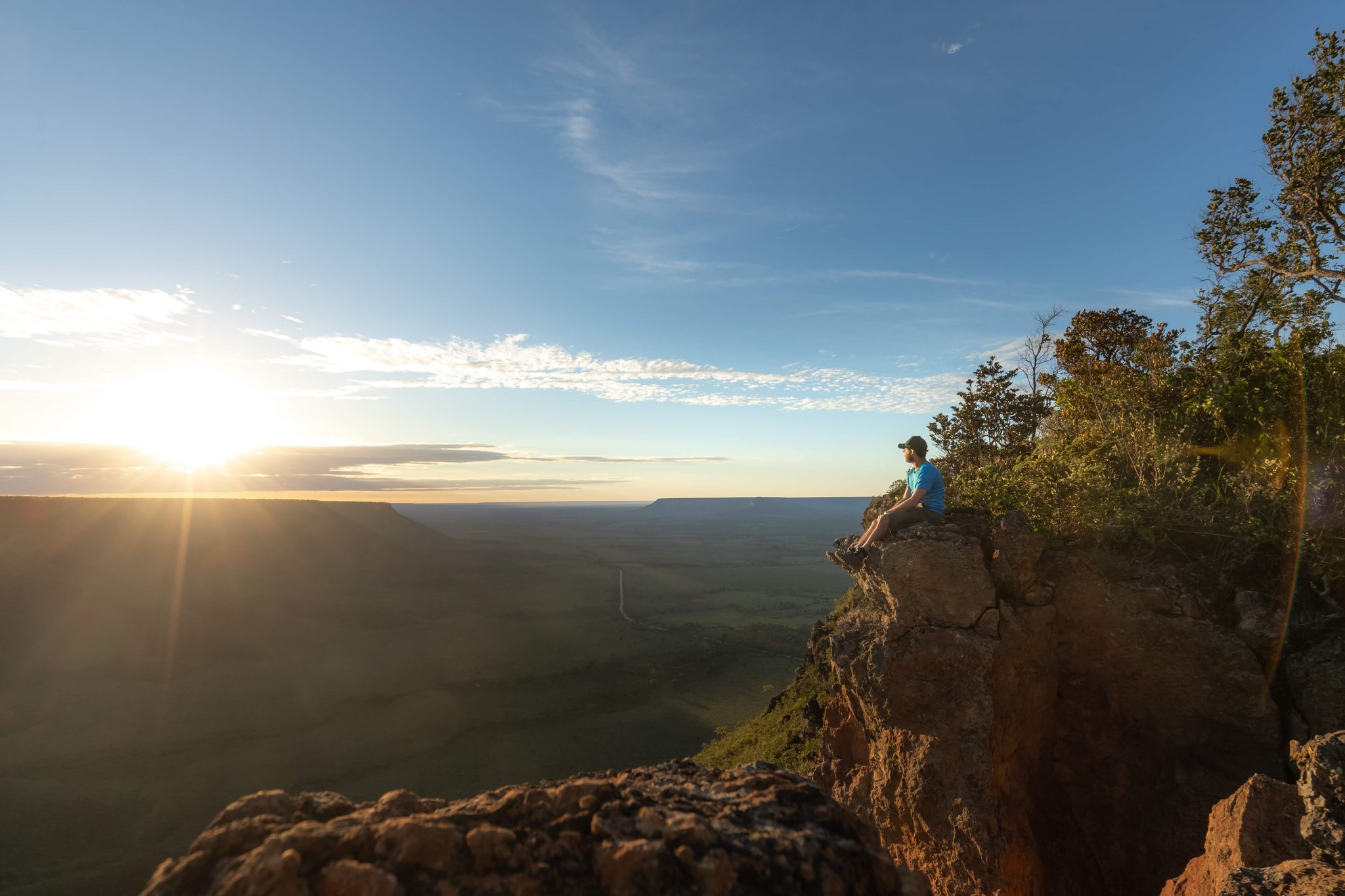 A lone traveler sits on the edge of a sandstone cliff during a Jalapão trekking expedition, gazing out over a vast green plateau bathed in golden sunrise light