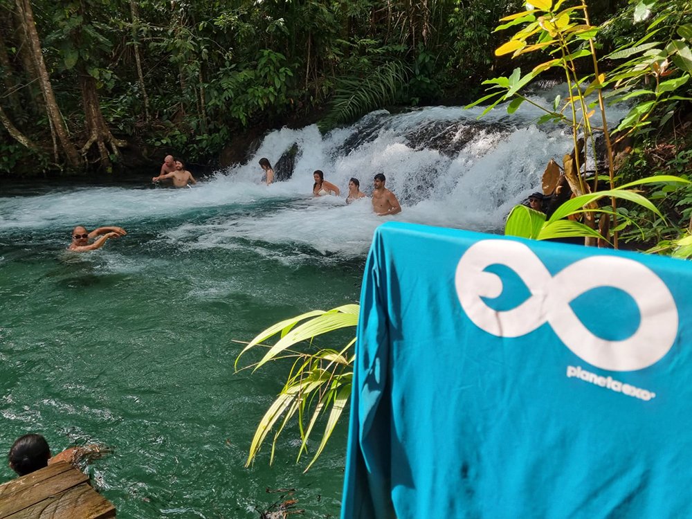 A group of travellers swimming and wading at the base of a cascading waterfall surrounded by dense tropical forest on a Jalapão trekking experience, with a PlanetaEXO branded towel in the foreground