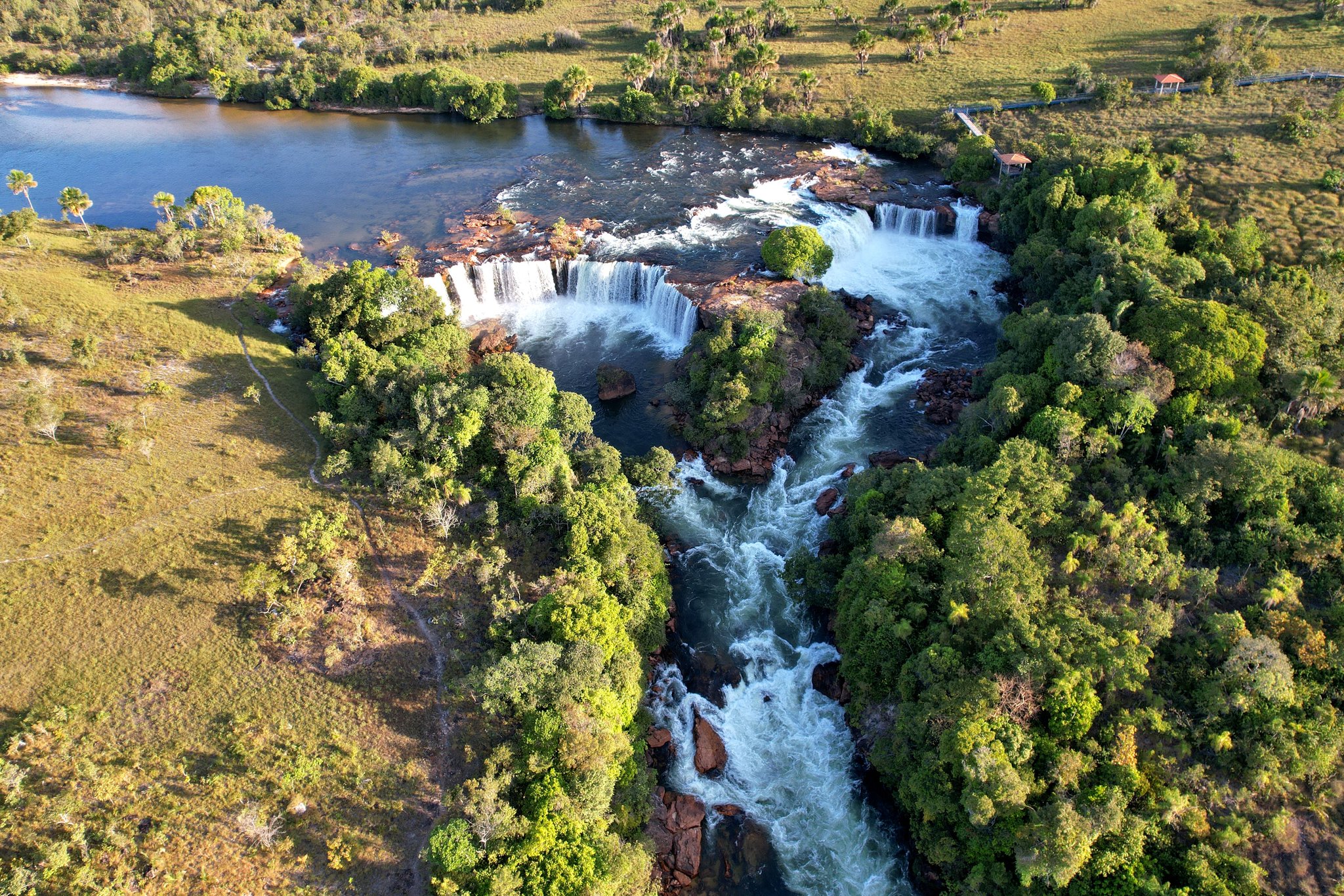 Three curtains of whitewater, one roaring descent — wild Brazil from above. Photo: Wirestock Aerial view of a wide multi-tiered waterfall on a jalapão trip, with white rapids churning through rock-lined channels, surrounded by cerrado savanna and a calm upstream pool