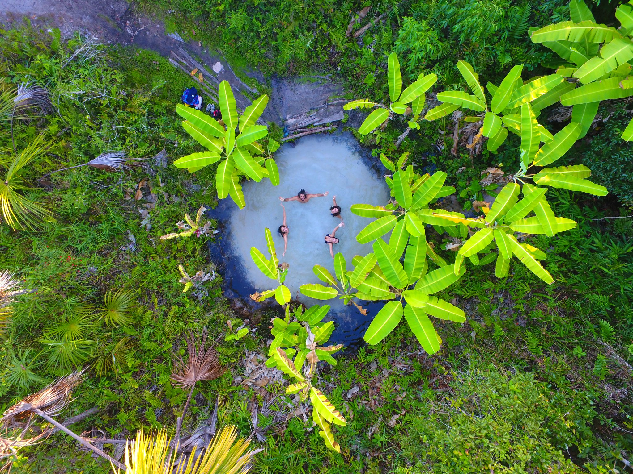 Four bodies, one hidden fervedouro — impossible to sink, impossible to forget. Aerial drone view of four swimmers floating in a small turquoise natural spring pool surrounded by lush tropical vegetation on a Jalapão trip