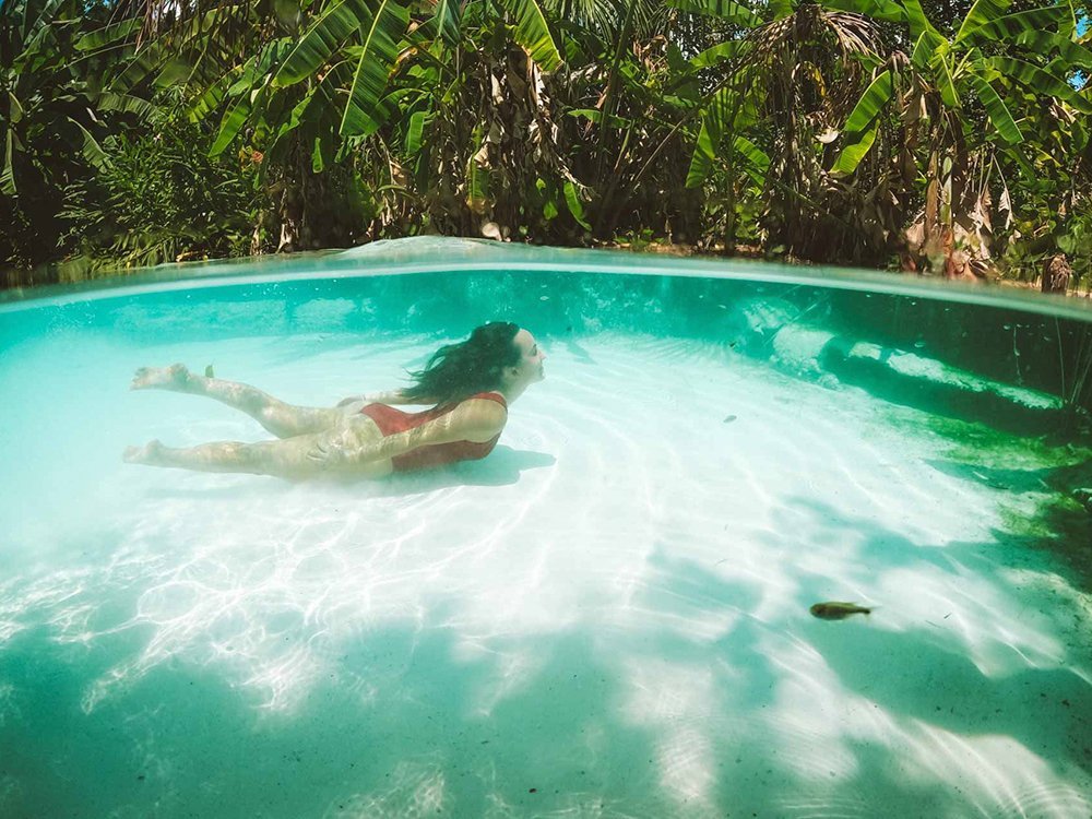 Weightless in turquoise — the fervedouros of Jalapão float you like nothing else. A woman in a red swimsuit glides beneath the crystal-clear turquoise surface of a fervedouro on a Jalapão trip, with a white sandy bottom and lush tropical foliage visible above the waterline