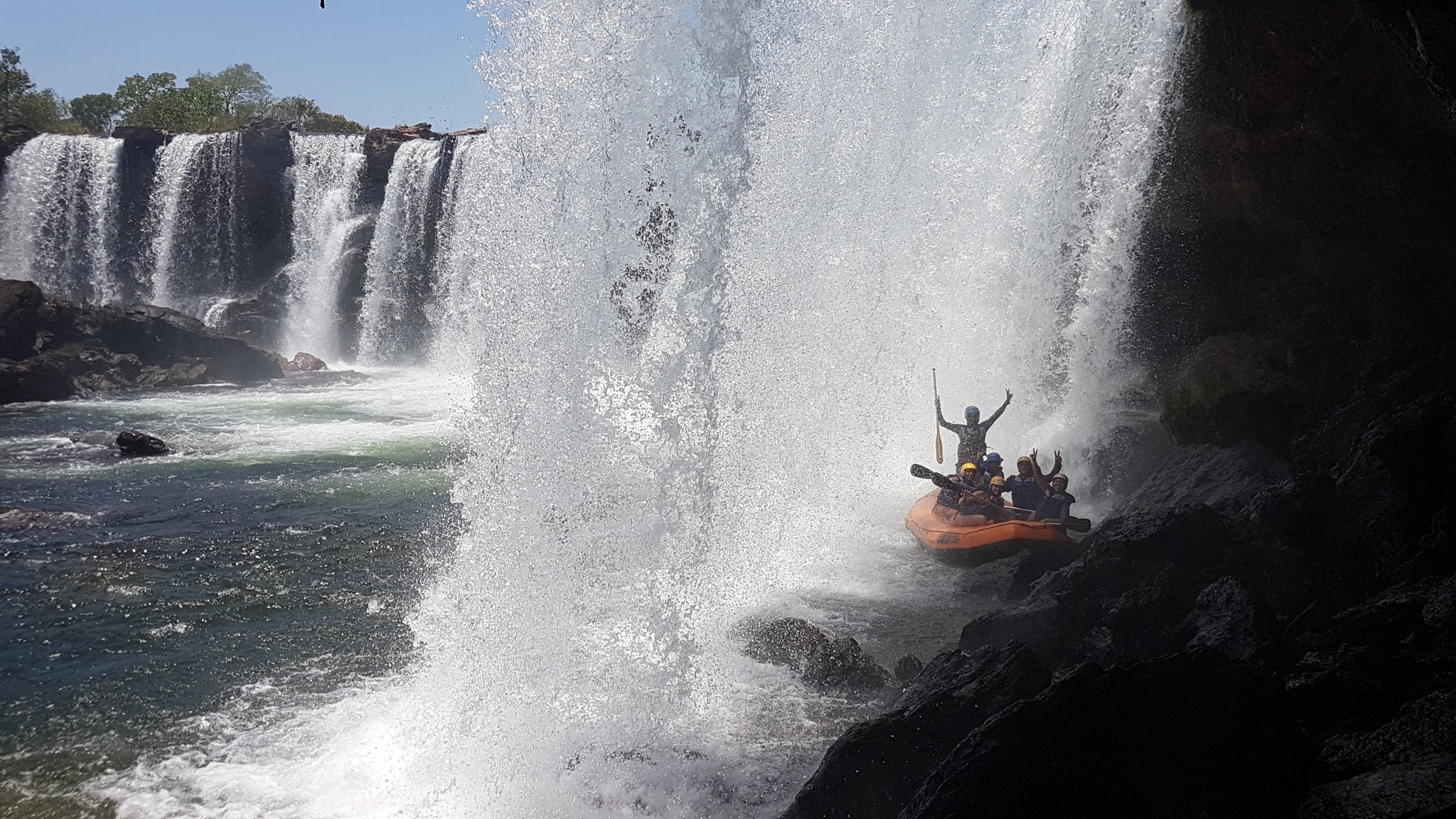 Soaked, screaming, and grinning — tucked behind the falls. An orange raft full of cheering passengers shelters beneath a thundering waterfall curtain on a jalapão trip, with a wide cascade spanning rocky cliffs in the background