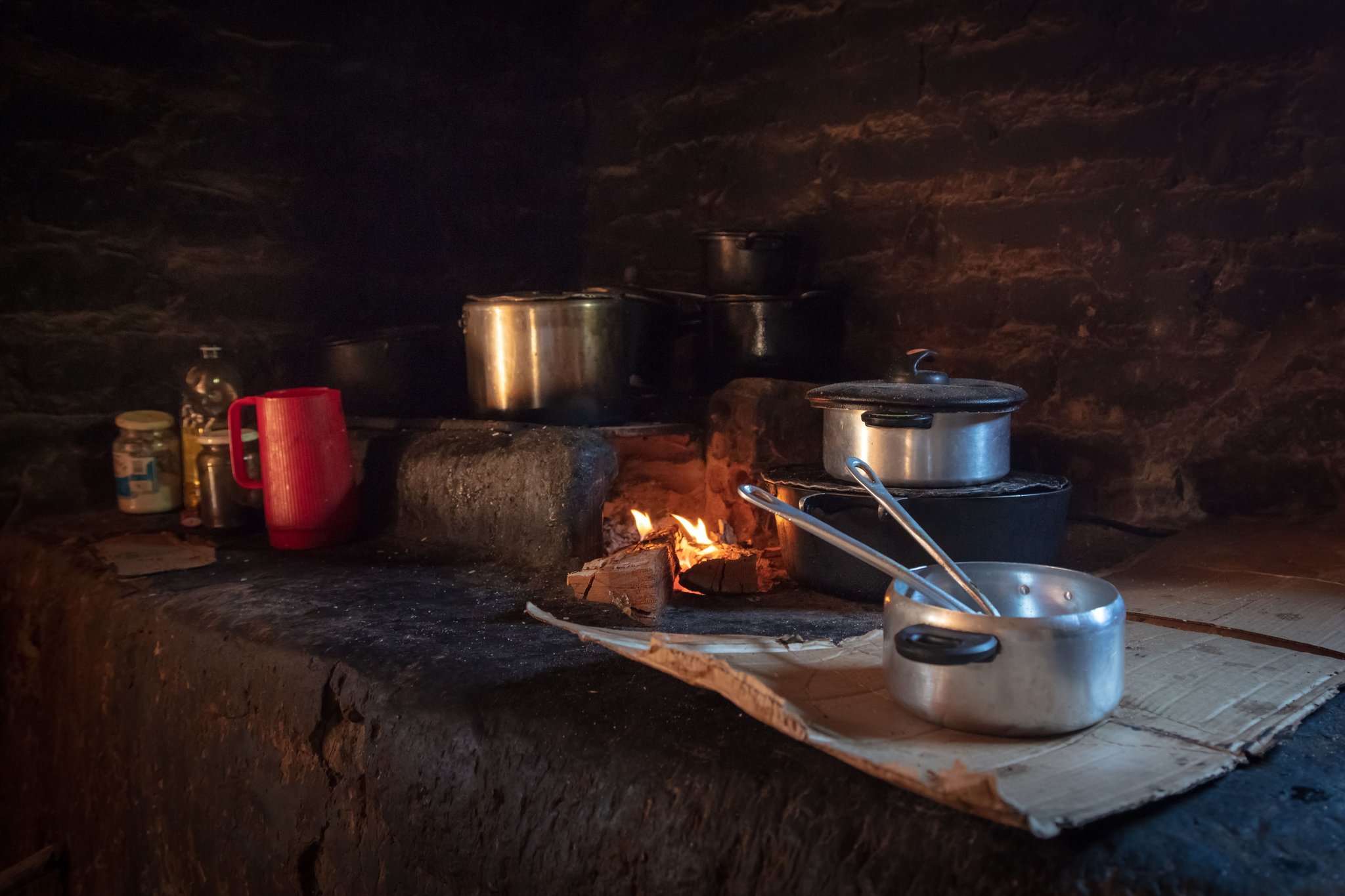 Multiple aluminium pots and pans simmering over an open wood fire on a rustic stone hearth, with a red mug and condiment jars nearby, in a dimly lit traditional kitchen on a jalapão trip