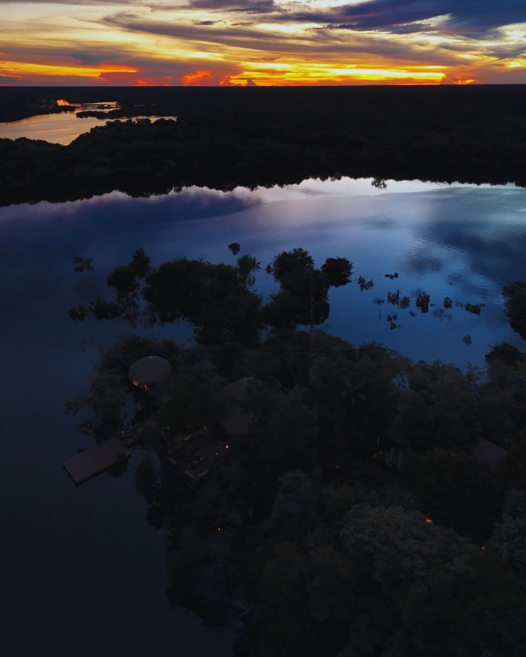 Aerial drone view of Juma Amazon Jungle Lodge nestled among flooded forest islands at dusk, with warm lodge lights glowing and a dramatic orange-and-gold sunset reflecting across a wide dark river
