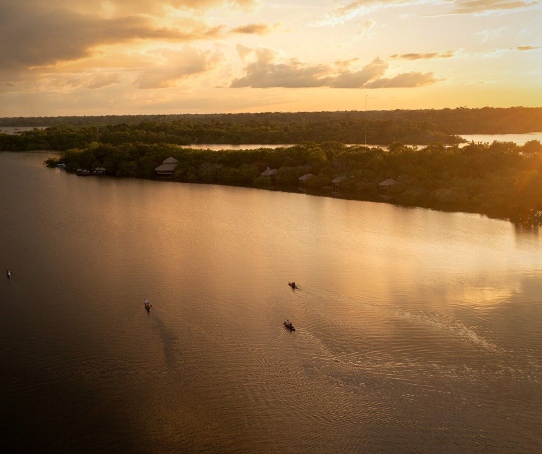 Golden hour from above — tiny canoes trace ripples across the river while the lodge waits quietly in the trees. Aerial view of small canoes gliding across a wide river at golden sunset, with the thatched bungalows of Juma Amazon Jungle Lodge nestled among dense rainforest on the far bank