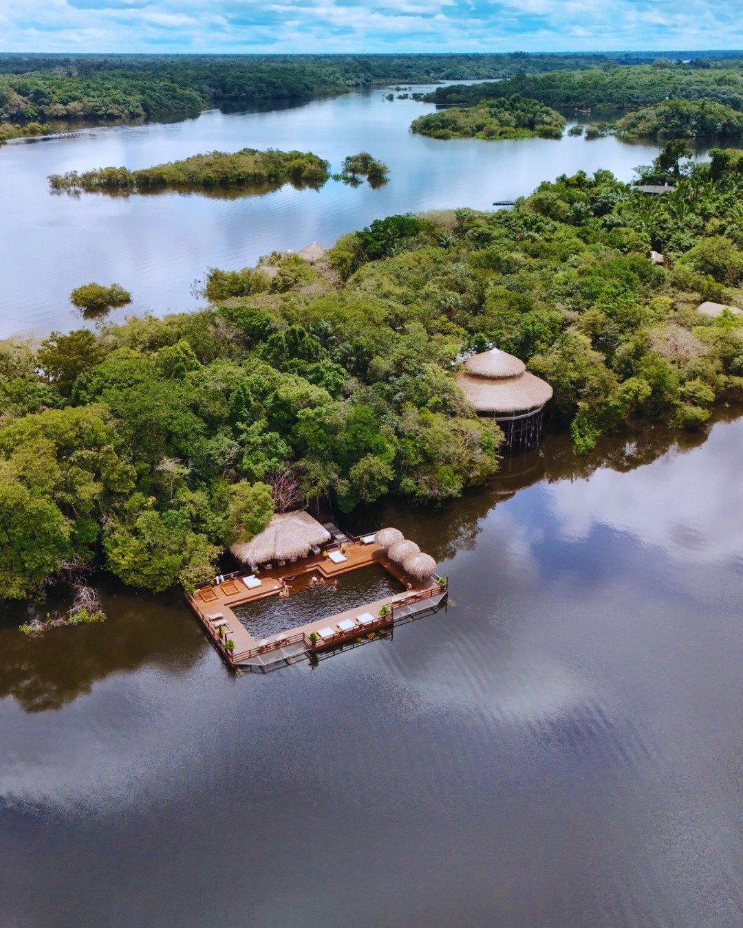 Floating above dark Amazonian waters, the lodge's open-air deck sits perfectly at the forest's edge. Aerial view of Juma Amazon Jungle Lodge showing a wooden floating deck with thatched-roof pavilions and a river pool, surrounded by flooded rainforest and dark Amazonian waters