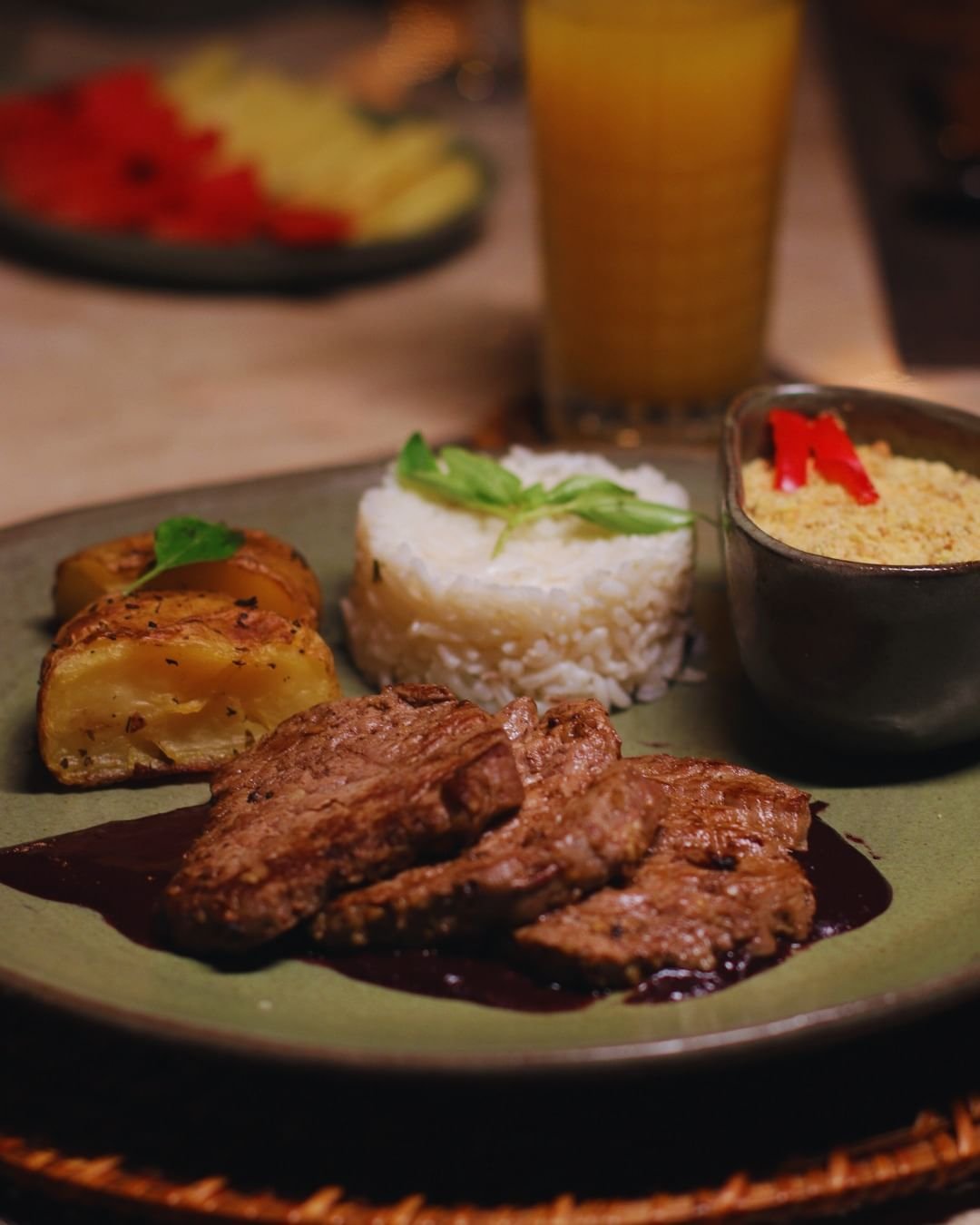Classic Brazilian flavours, freshly plated after a day deep in the rainforest. A plated dinner at Juma Amazon Jungle Lodge featuring sliced grilled beef in dark sauce, a moulded rice garnished with fresh herbs, herb-roasted potato wedges, and a small bowl of farofa topped with red pepper strips, served alongside a glass of orange juice