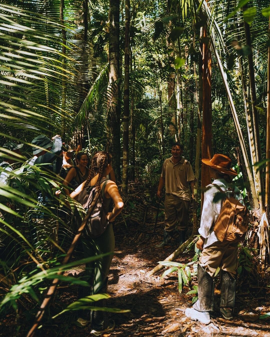 Deep in the forest, laughter echoes between the palms. A small group of smiling travellers follows a local guide along a narrow trail through dense tropical rainforest at Juma Amazon Jungle Lodge, with palm fronds and tall trees surrounding the path
