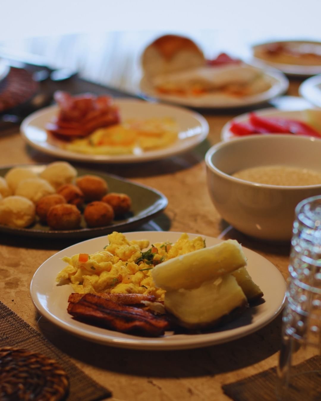 Fuel up before the forest: scrambled eggs, cassava, and all the trimmings. A generous breakfast spread at Juma Amazon Jungle Lodge, with scrambled eggs, crispy bacon strips, boiled cassava, cured meats, fresh red pepper, fried snacks, rolls and a bowl of porridge arranged across a wooden table