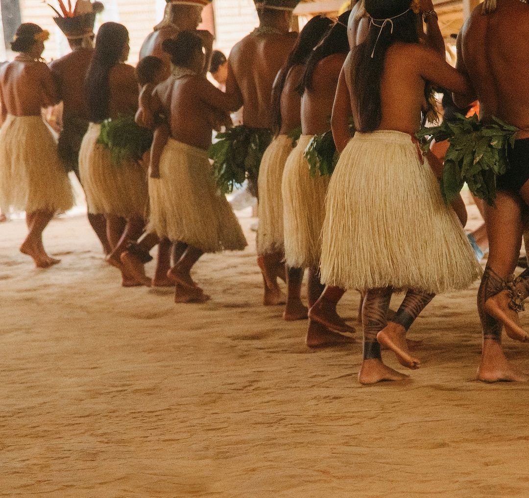 Straw skirts swaying in unison — living tradition, passed down through generations. A line of indigenous dancers wearing woven straw skirts and fresh leaf adornments move barefoot across a sand floor during a traditional ceremony at the Juma Amazon Jungle Lodge community visit