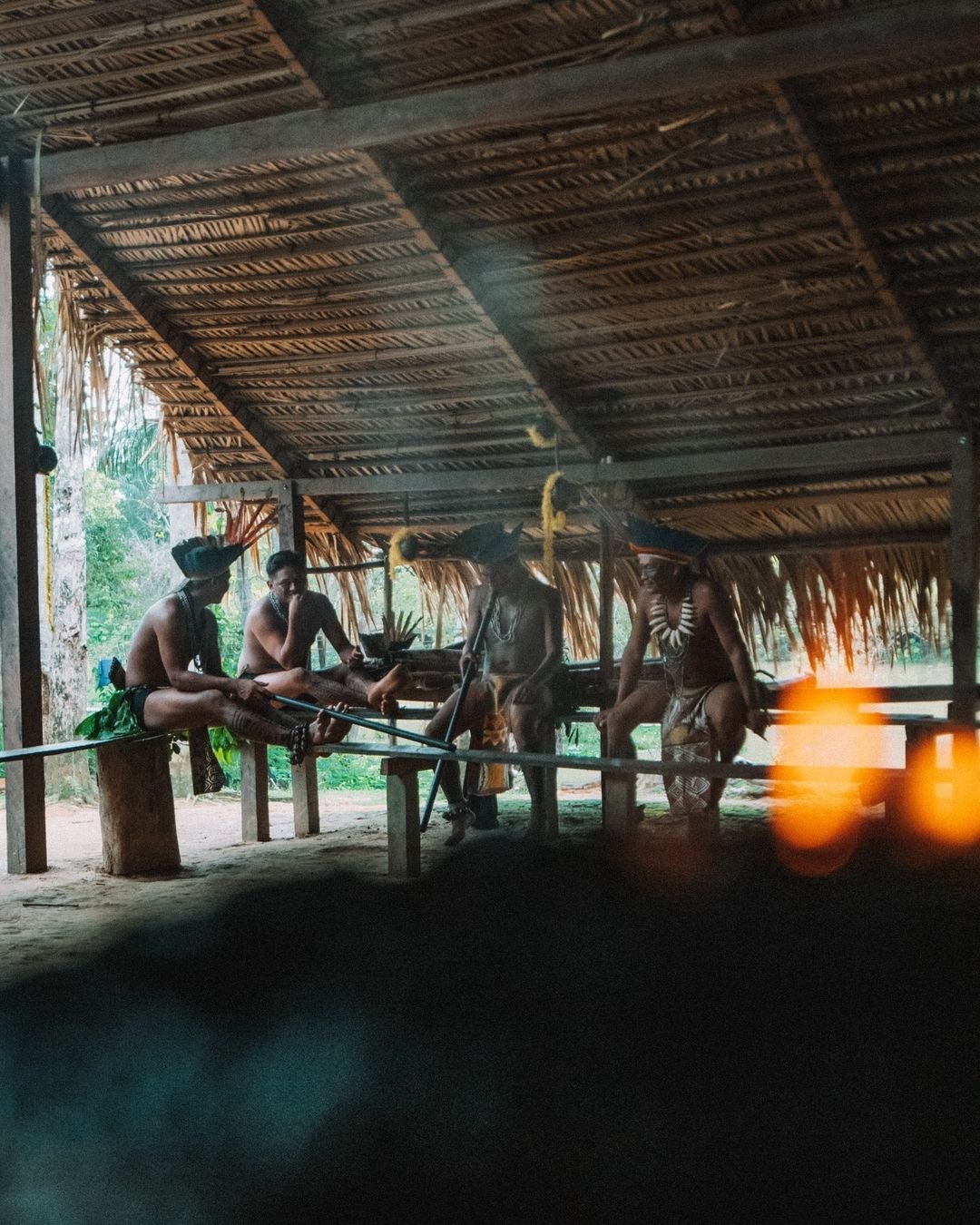 Smoke, stillness, ceremony — a glimpse into living Amazonian tradition. Indigenous community members in traditional body paint, headdresses and beaded necklaces gathered under a thatched-roof open-air structure at Juma Amazon Jungle Lodge, long blowpipes resting across wooden benches